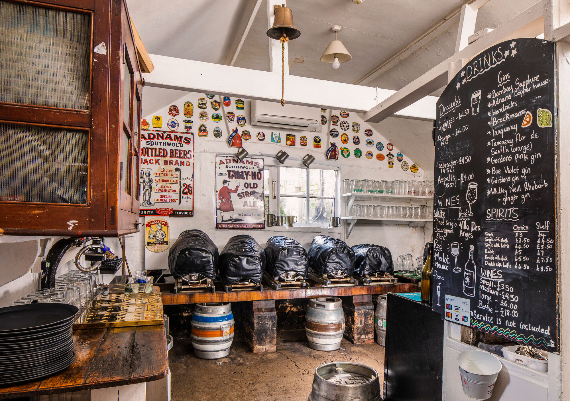 A view of a taproom in a public house, with barrels arranged along a low sturdy wooden counter and glasses on shelves, in the foreground is a chalk board sign with prices.