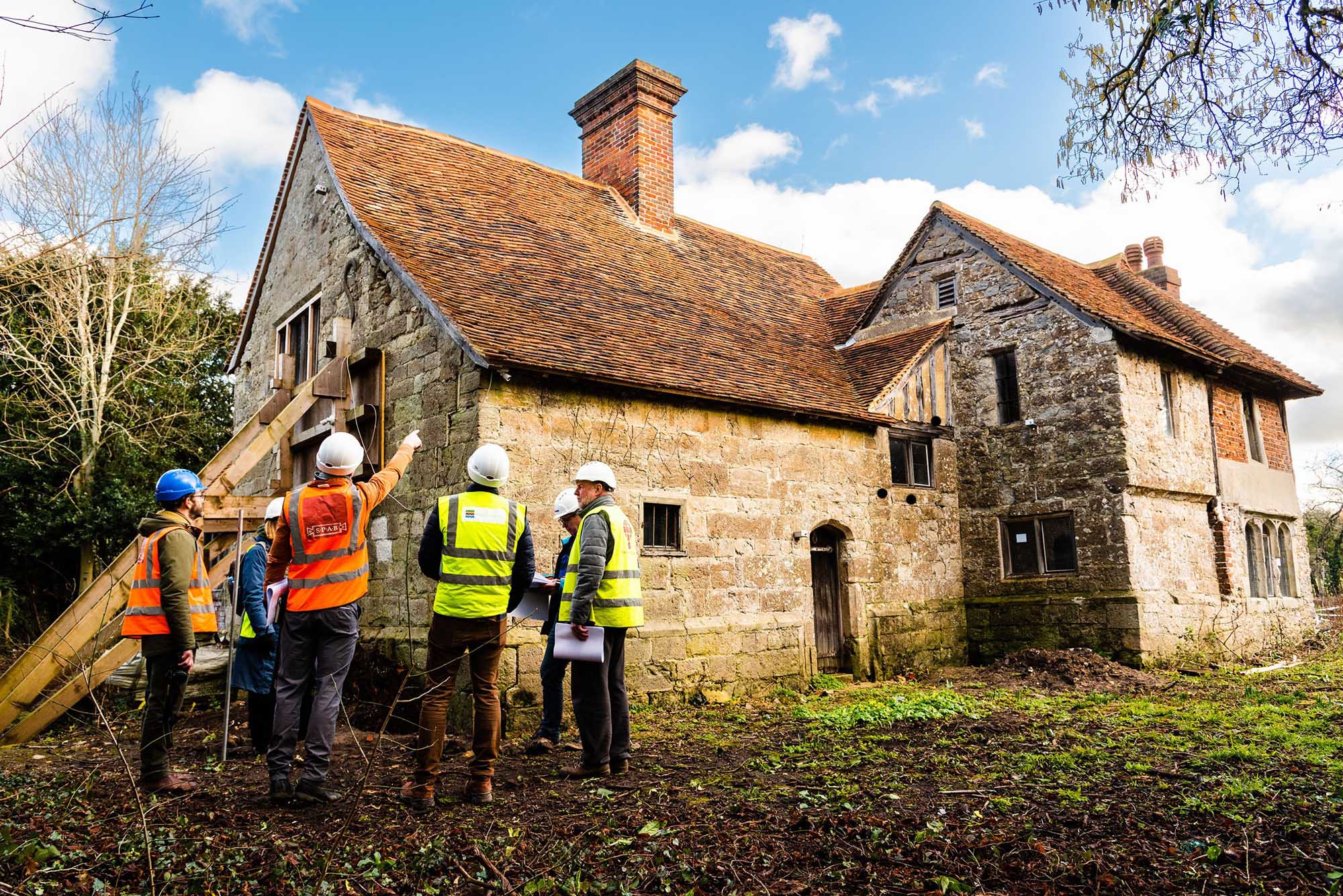 A group of people in hard hats and high-vis jackets look at an old stone building.