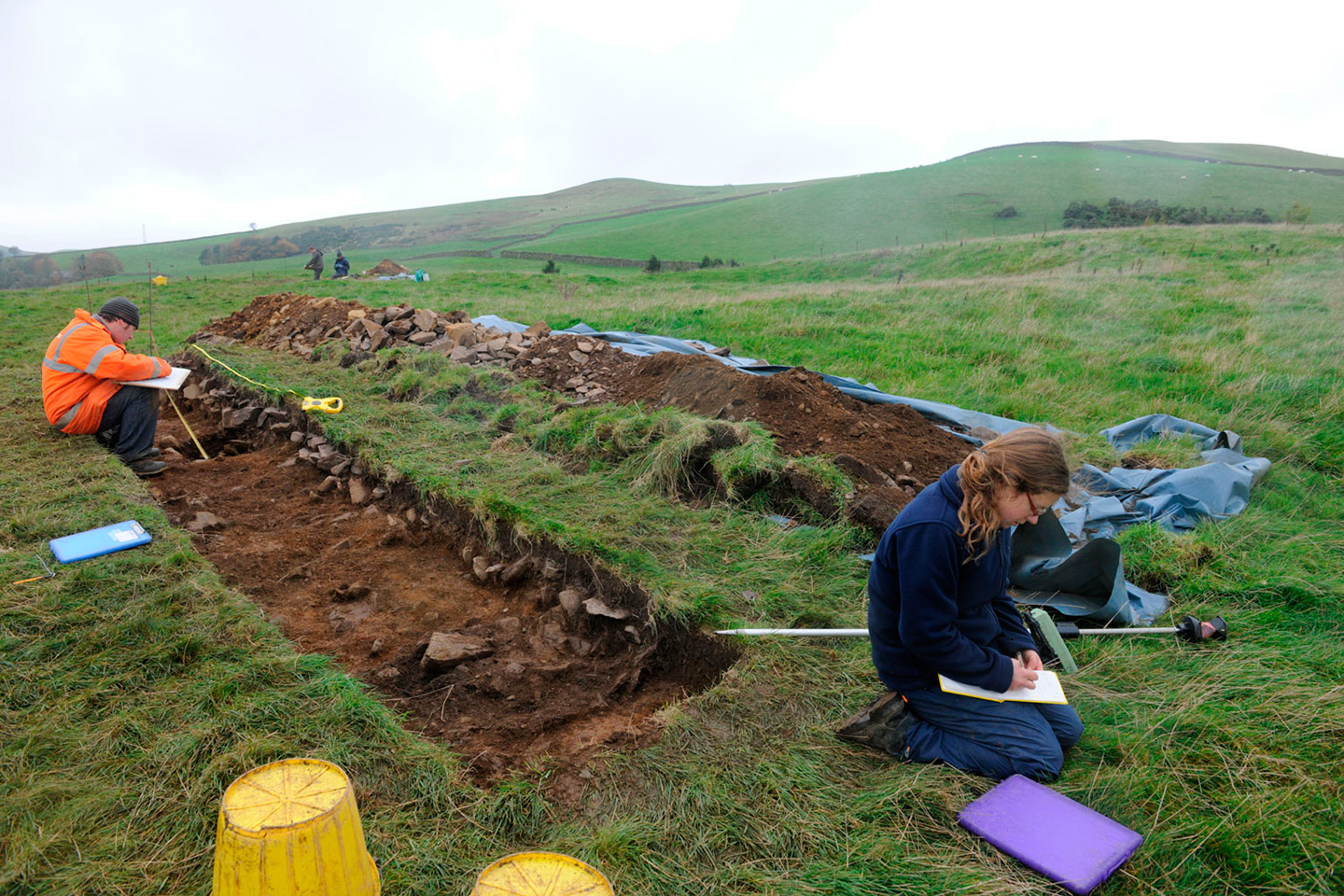 An open excavation trench reveals the remains of a collapsed stone bank. Archaeologists are working in the trench.