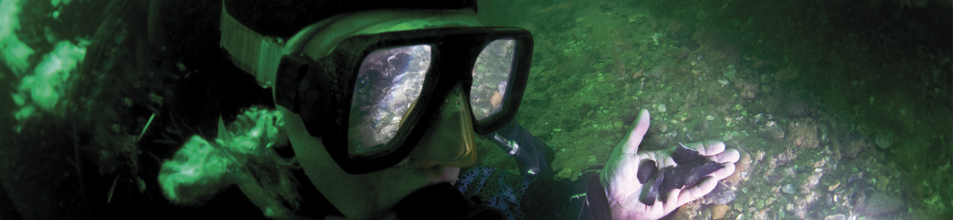 Close up photograph of diver’s face looking at hand holding flints from prehistoric site underwater