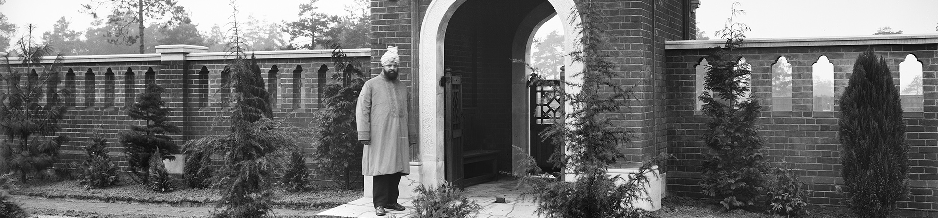 A man wearing a turban stands in front of the Indian inspired gateway of the Muslim Burial Ground, Horsell Common, Surrey. A wall pierced with Indian style tracery surrounds the cemetery.