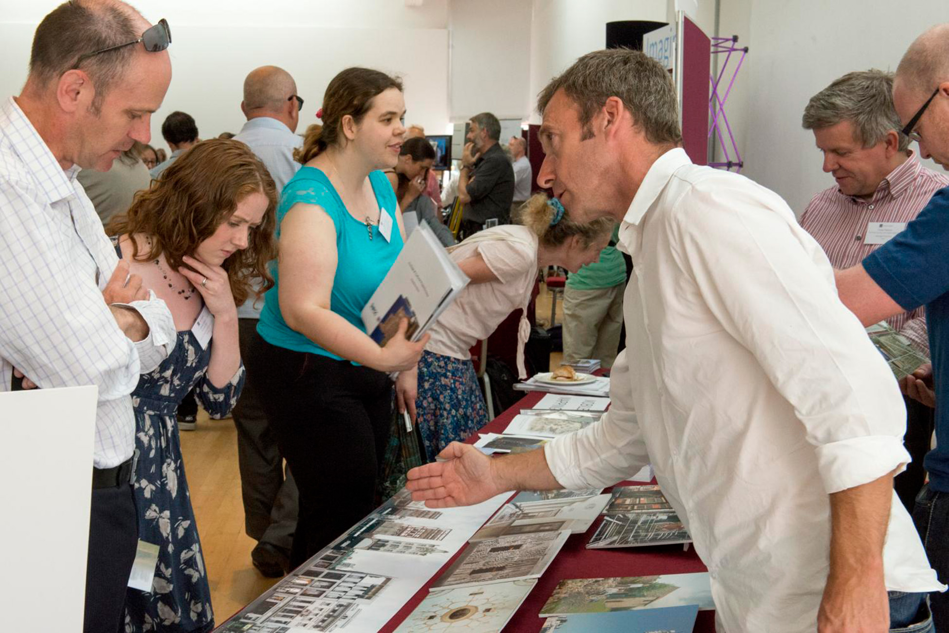 A photo of a busy conference exhibition stand surrounded by people. A man in a white shirt is gesturing to a man and woman looking at a display in the foreground.