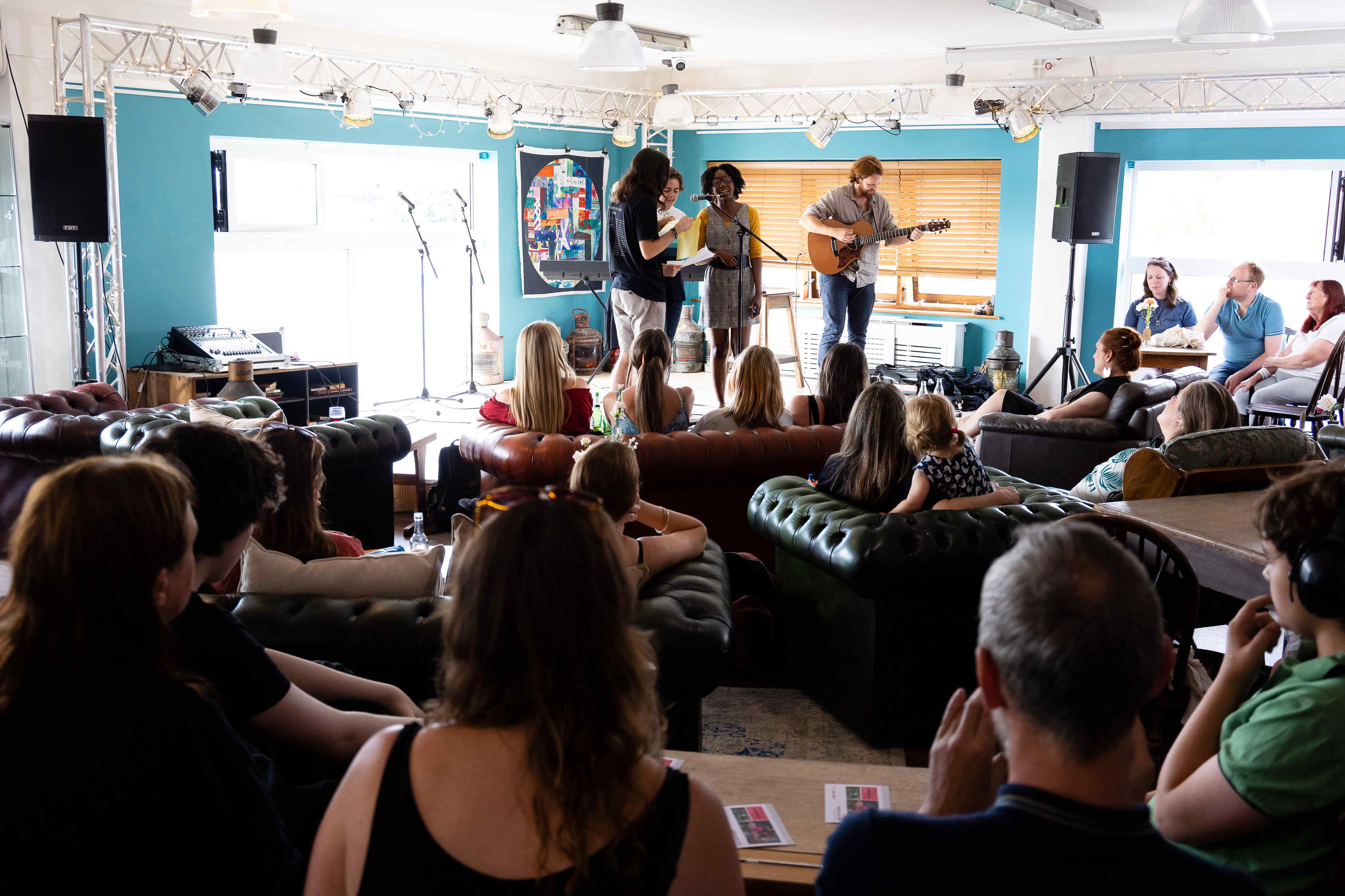 Performance in Chatham, musicians on stage singing into a microphone whilst a guitar is played. Audience sit in sofas facing the stage.