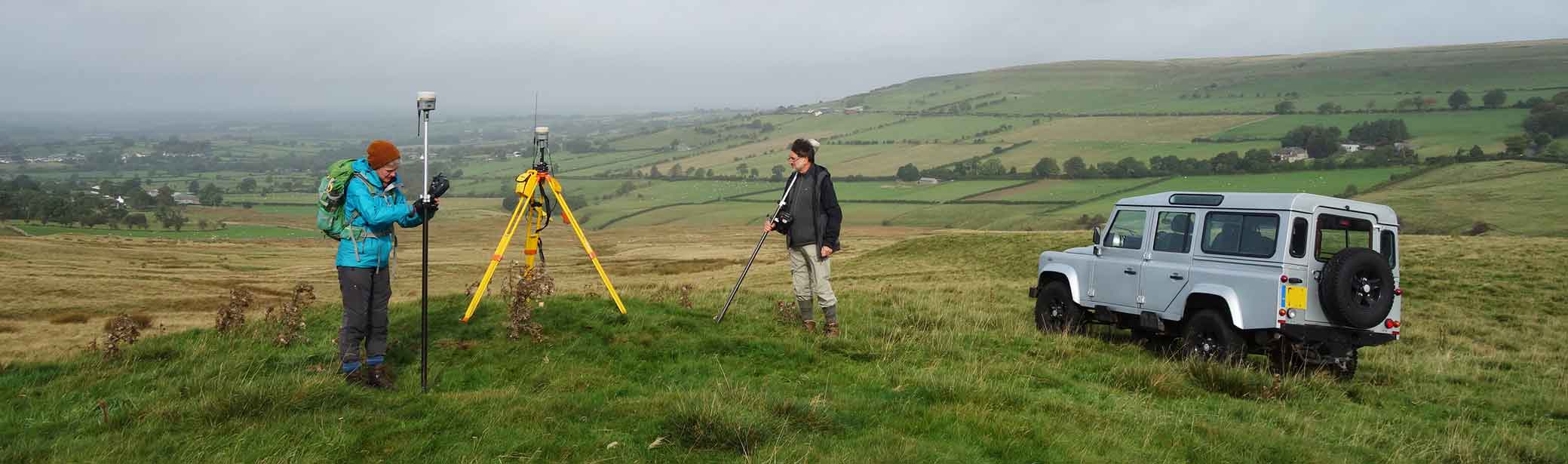 Archaeologists using surveying equipment with a a fell landscape in the background.