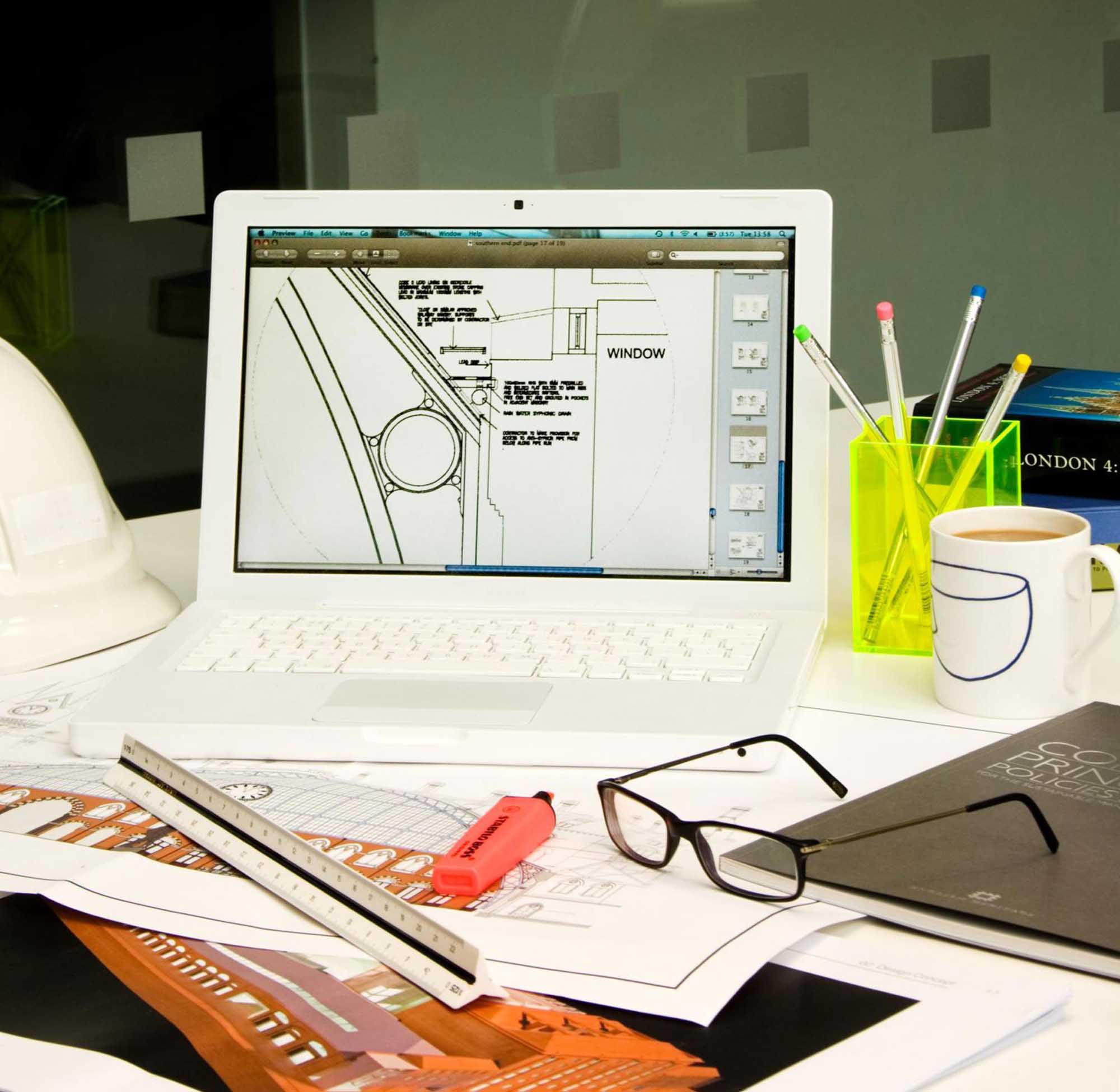 Detail of office environment including building plans on a laptop screen and a hard hat on the desk next to it.