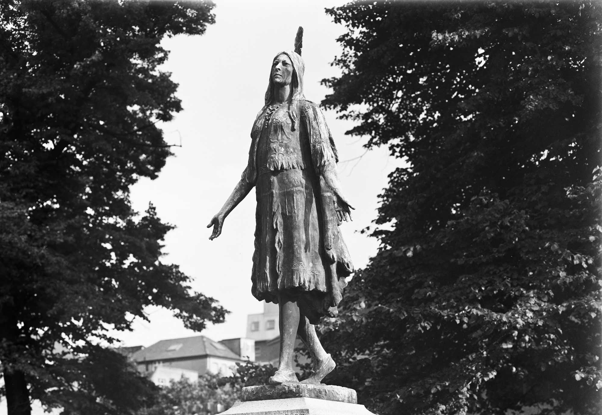 A black and white image of a Pocahontas statue with trees and buildings in the background