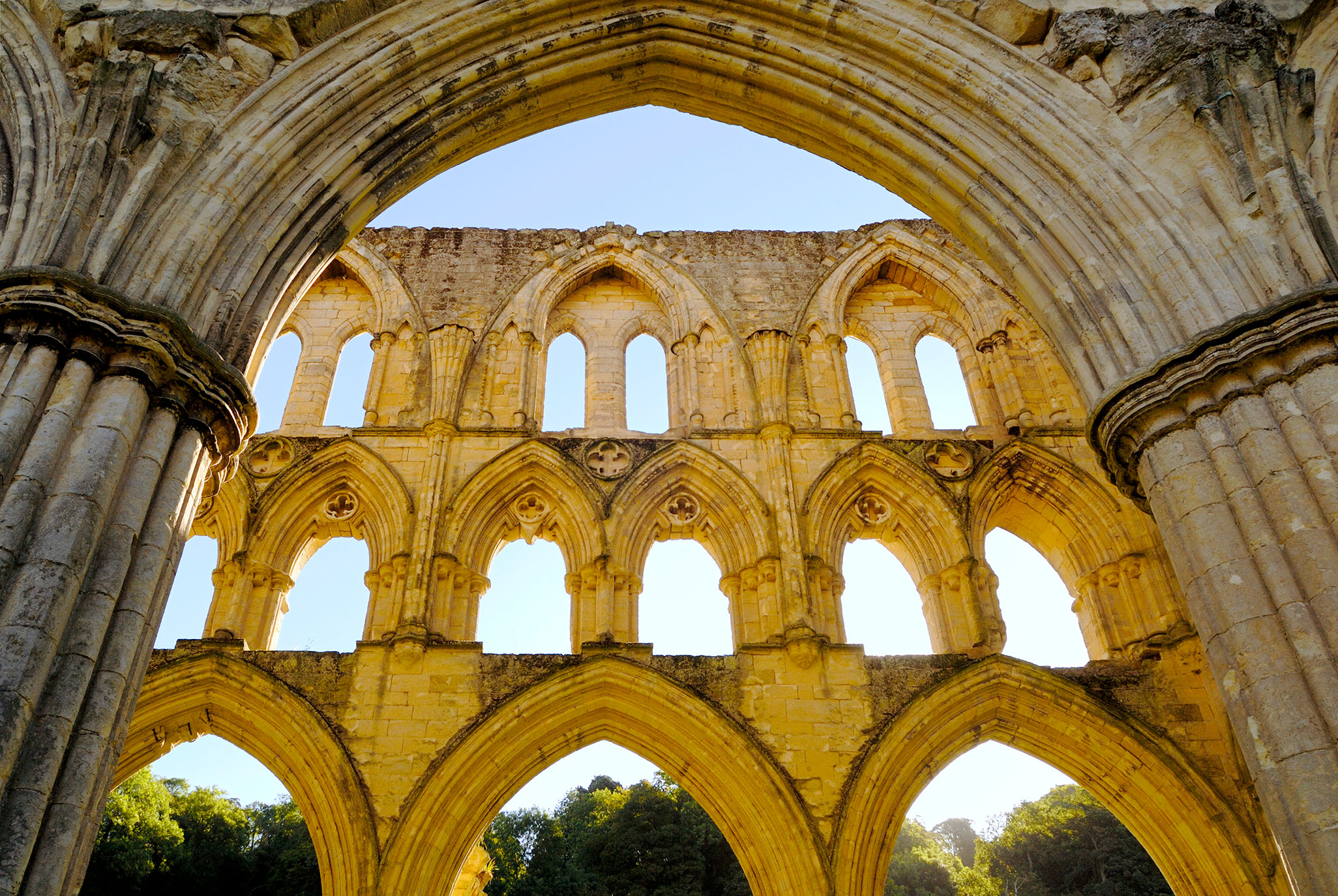 The ruins of an ancient stone structure, featuring intricate arches and windows, framed by a clear blue sky and lush greenery in the background