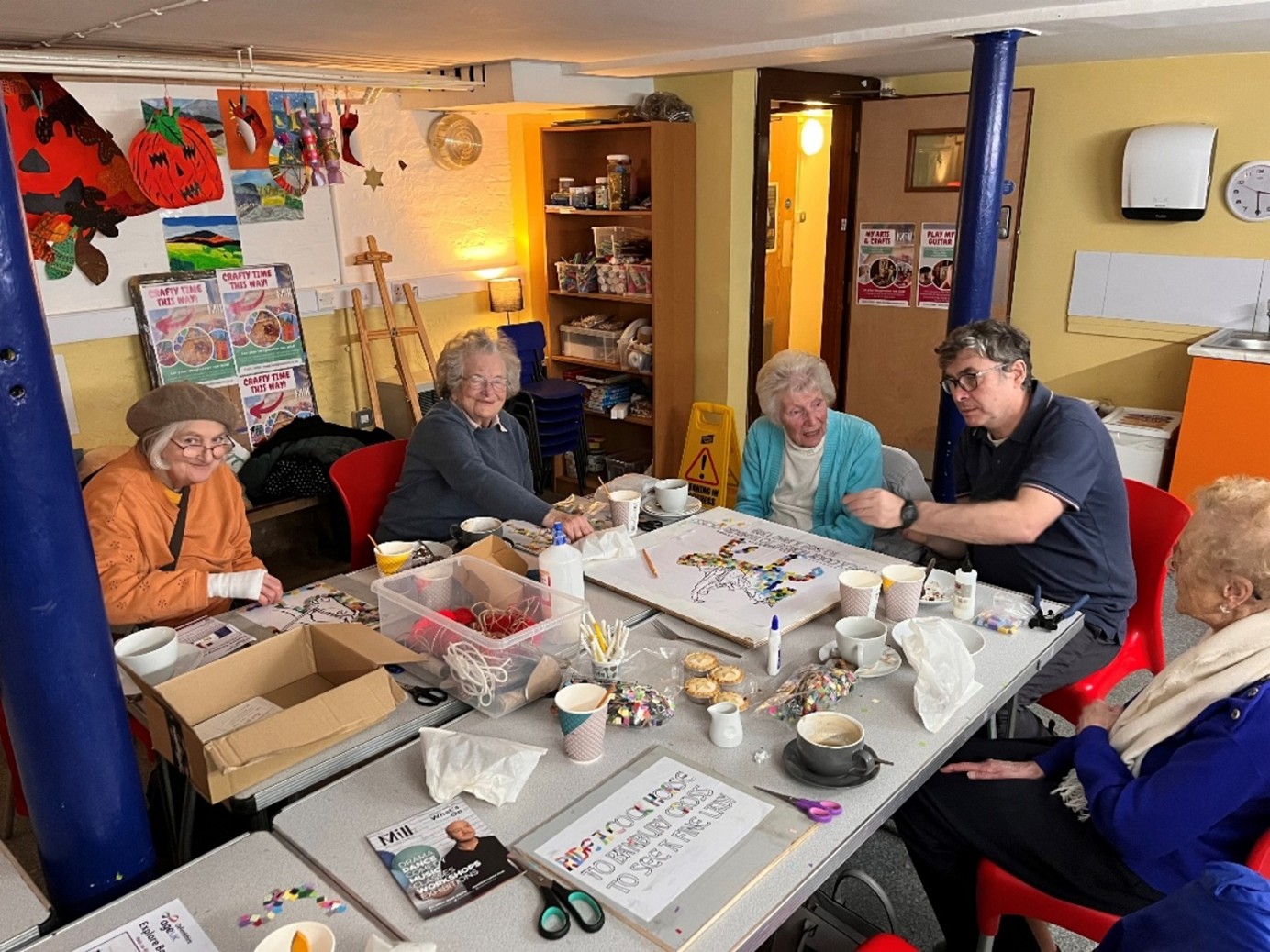 A group of elderly ladies and a younger man are sitting around a table covered with artwork materials.