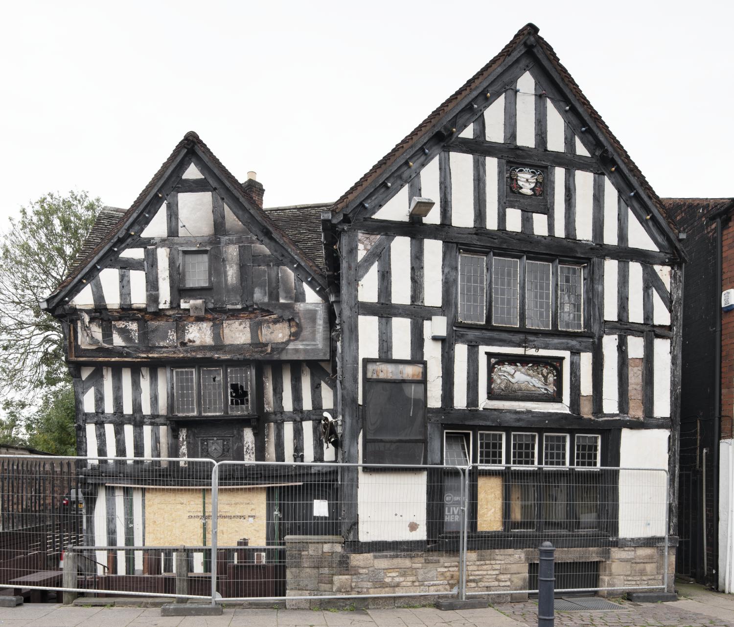 A timber-framed building behind a metal safety fence with fire damage visible on the upper floors and boarded up windows on the ground floor.
