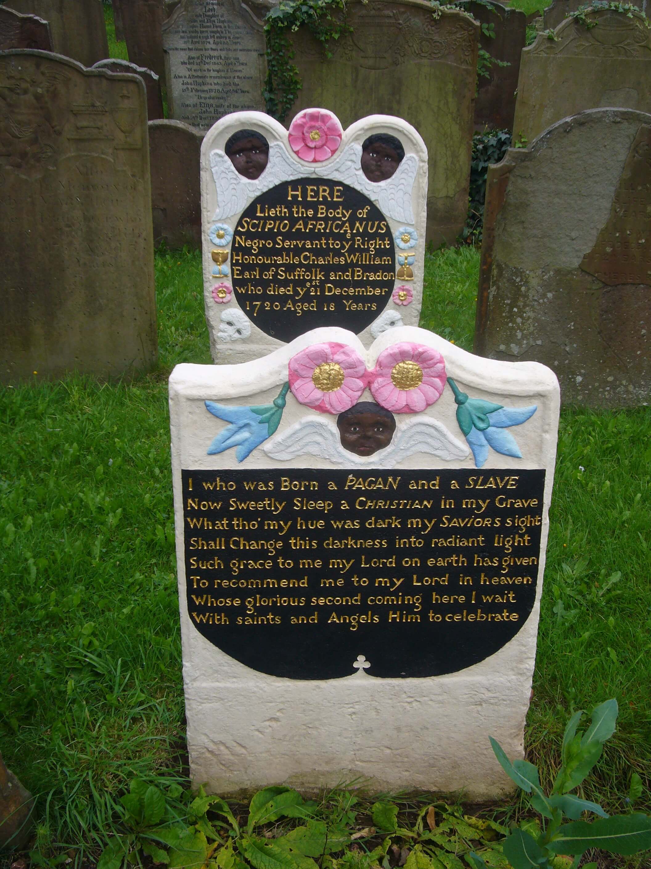 Headstone and footstone, with shaped tops. The freshly painted memorial stands out amongst unpainted stones. Carving includes winged Black cherubs, and flowers. The headstone carries the details of the subject’s life; the footstone carries a verse.