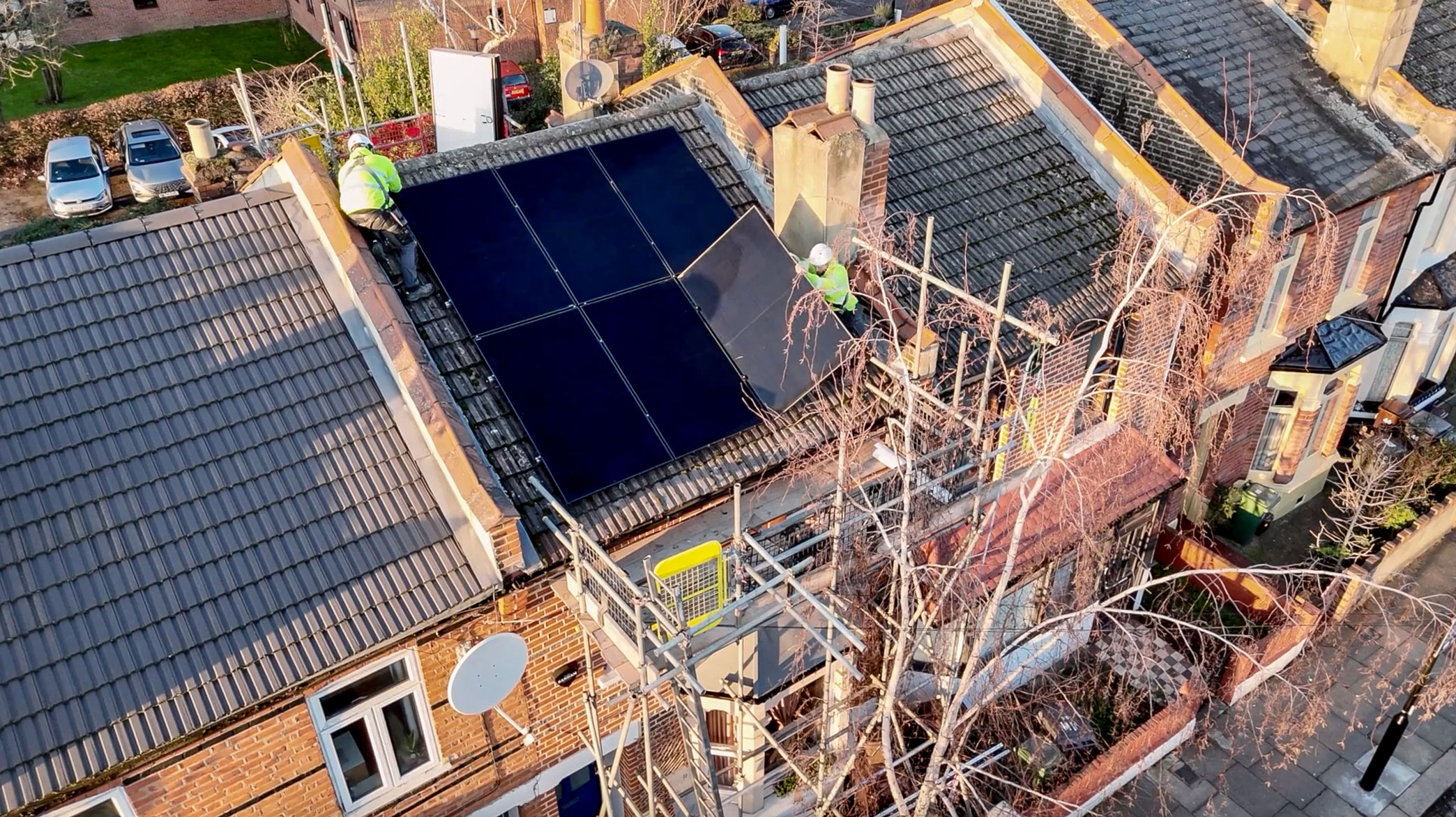 Workers in hi-vis gear are installing solar panels on the roof of a typical Victorian terraced house.