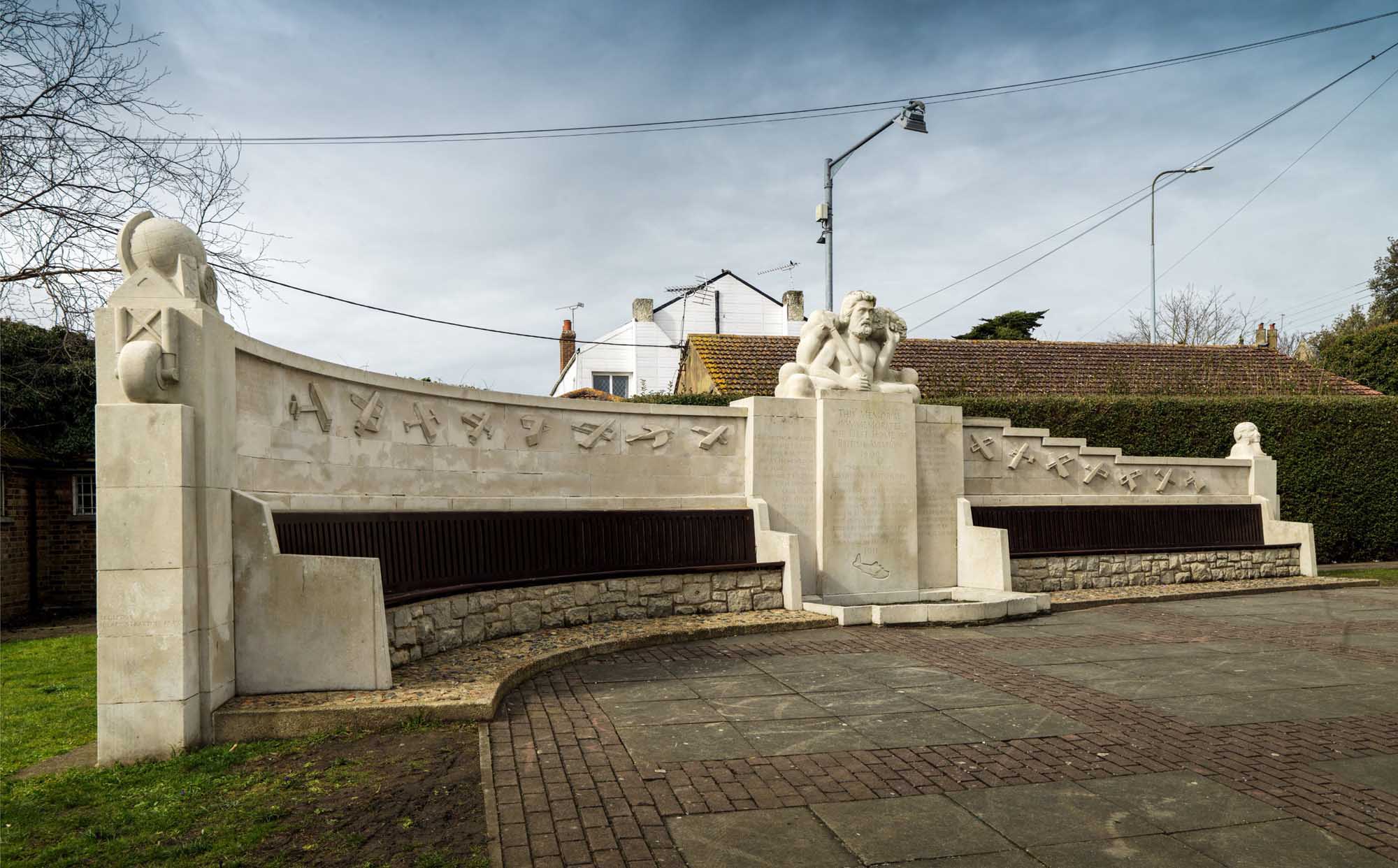 Figure of Zeus supported by plinth with wall. Plinth with projecting centre with dedication and recessed sides with names. Incised aircraft depicted below dedication. Wall depicting relief of eighteen early aircrafts in flight extends from rear of plinth. Whole set in paved area. 