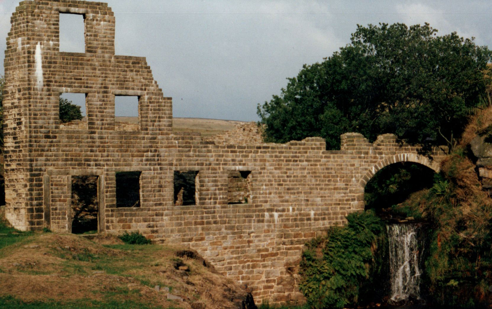The stone facade of a ruined textile mill, featuring an arch over a waterfall.