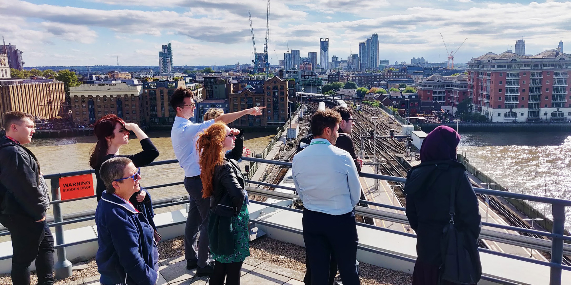 A group of people stood on a city rooftop and looking off in the direction of where the instructor is pointing. Skyscrapers, buildings, train tracks and a river are visible in the background.