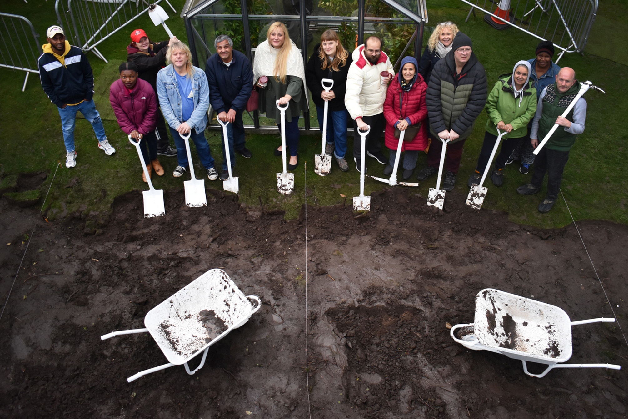 A group of people stood in a community garden smiling while holding muddy white spades, with white wheelbarrows in the foreground.