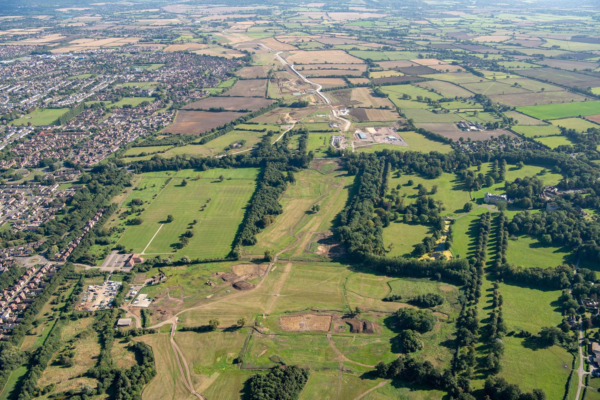 An aerial view of construction sites amid fields, with rows of houses in the background.