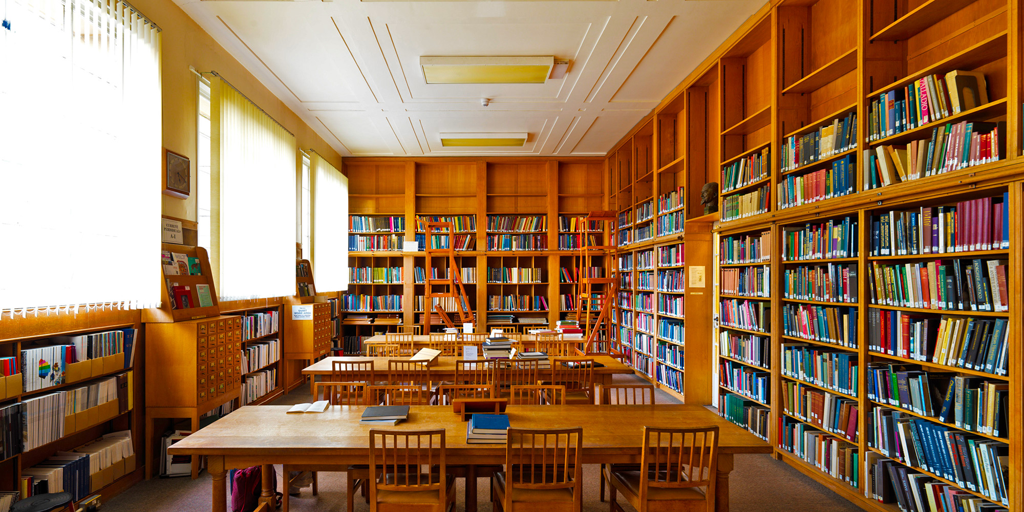 An art deco orange pine-panelled room filled with bookshelves, with communal reading tables and warm light coming from shaded windows. 
