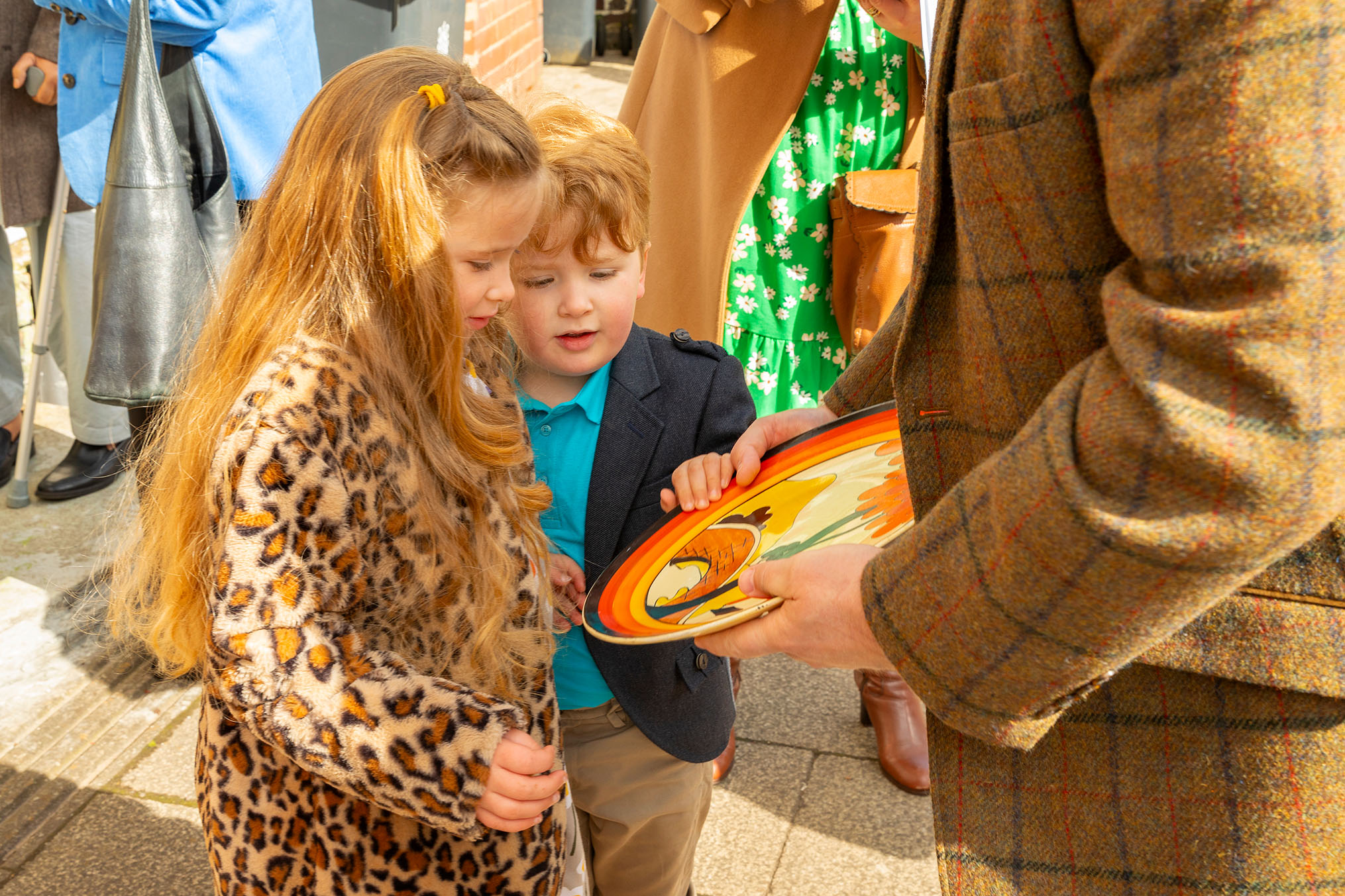 A close-up photograph of a young boy and girl looking at a colorful ceramic plate held by an older figure in a brown tartan jacket