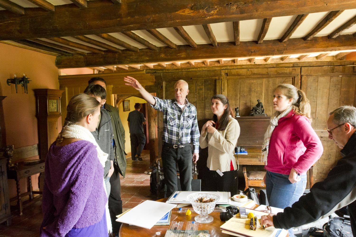 A group of six people stand in a room with wooden beams. A man in a checkered shirt gestures while others listen and look on. Papers and equipment are on the table in front of them.