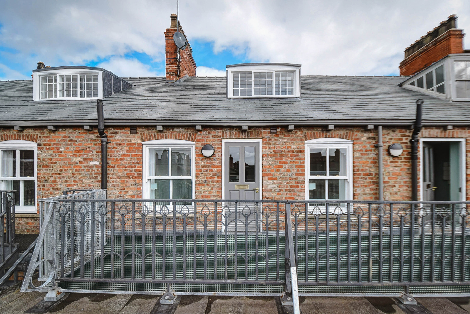 Exterior photo of 2 apartments on the first floor of an old brick building. In the foreground are railings and a walkway on a flat roof area.