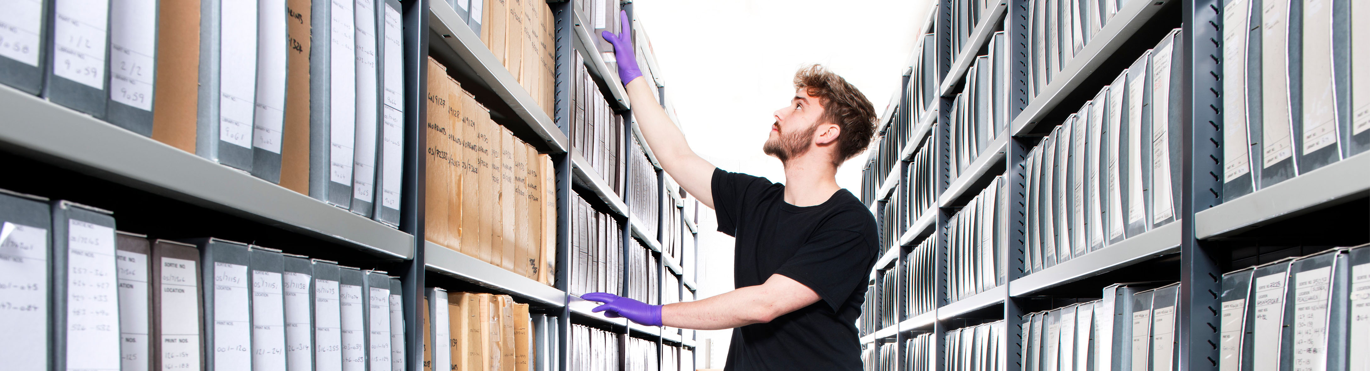 Colour photograph of a man wearing a black t-shirt and purple gloves, standing in an aisle between rows of shelving. He reaches up to remove or replace a box from the top shelf of the left-hand side of the shelving. 