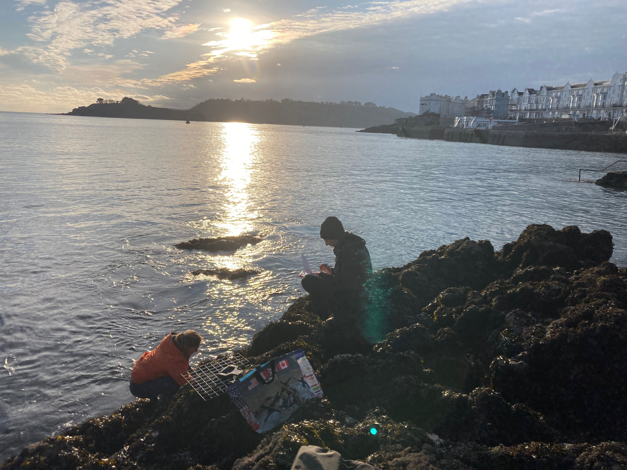 A man and a woman recording a rocky foreshore with the sea and coastal buildings in the background.