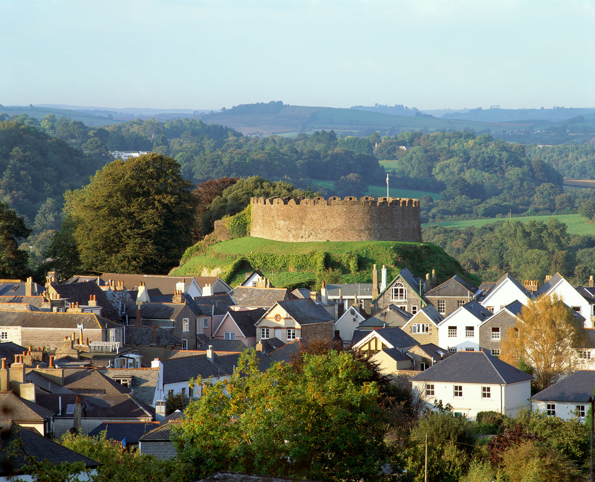 In the foreground is the pitched roofs of local houses, whilst in the background is Totnes Castle on a small hill, an example of a Norman motte and bailey castle. Rolling hills and woodlands can be seen in the background. Photo is taken in late spring or early summer.