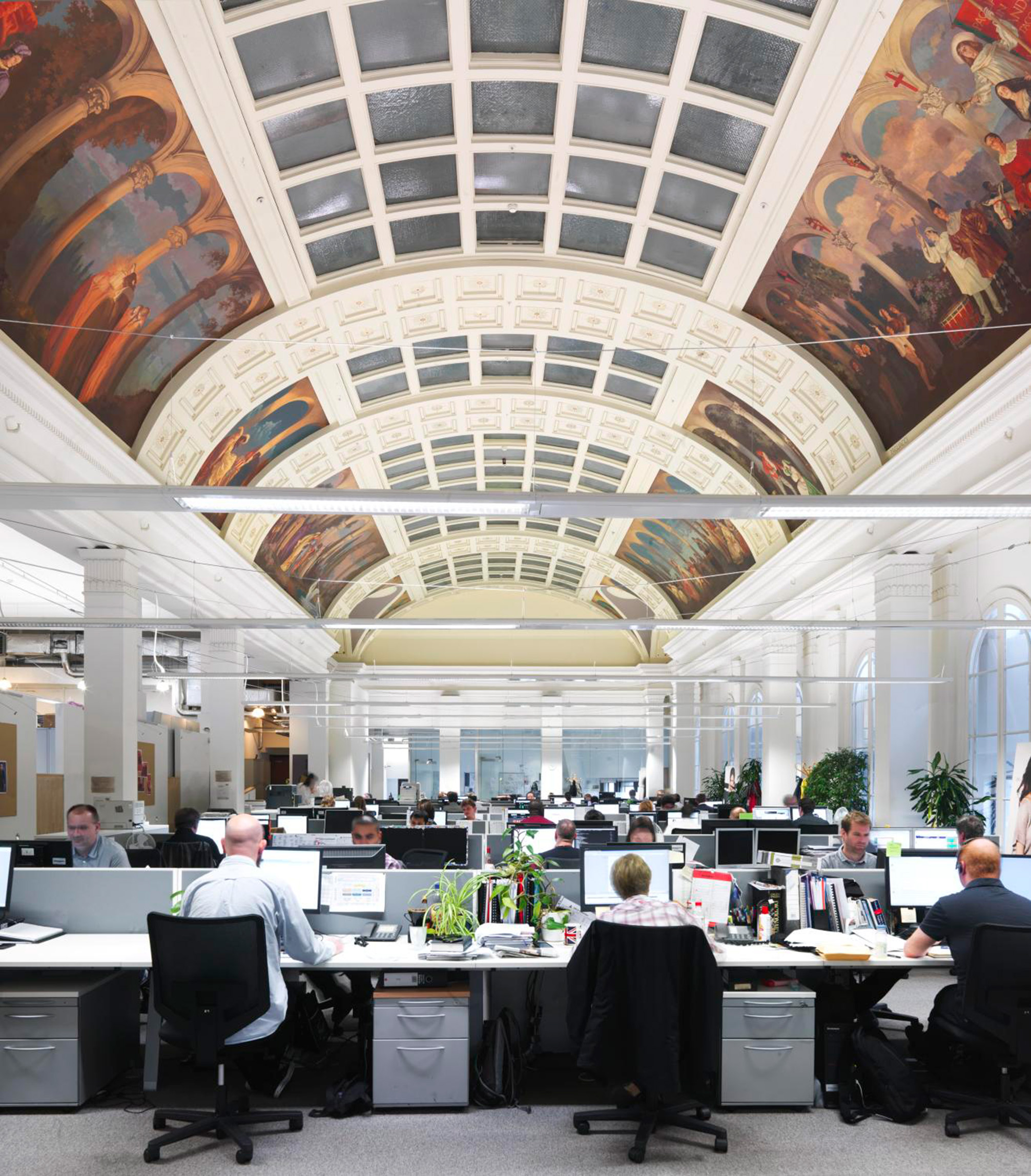 Many people working at computer desks in a well-lit office with an arched ceiling with classical painted frescoes and windows at the top of the ceiling.