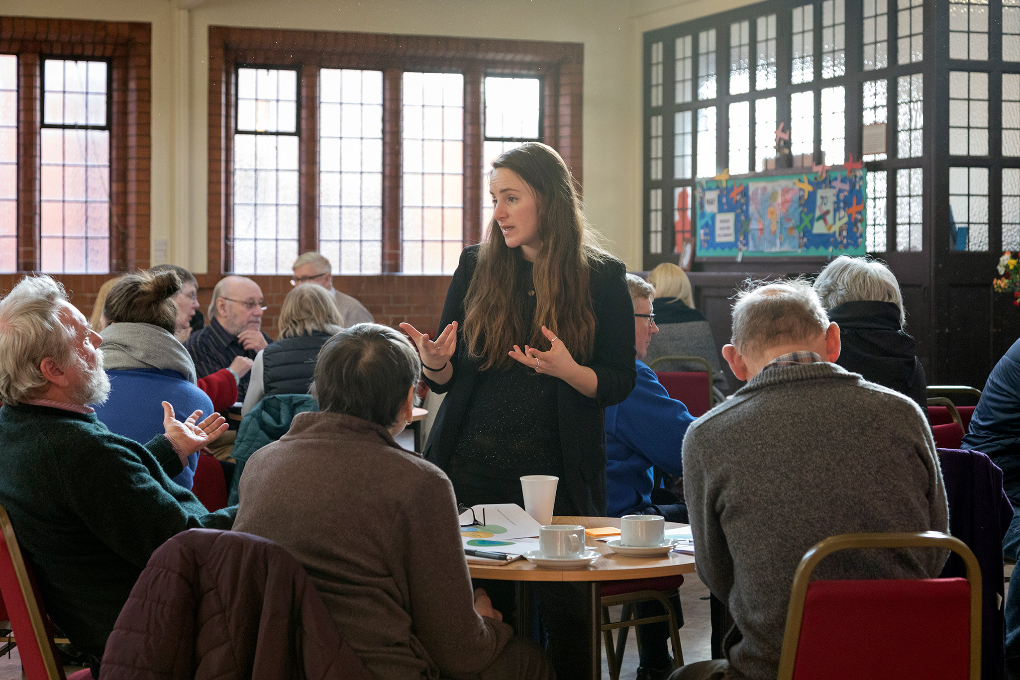 A person with long brown hair stands and gestures while speaking to seated elderly people in a community hall with tea and coffee cups.