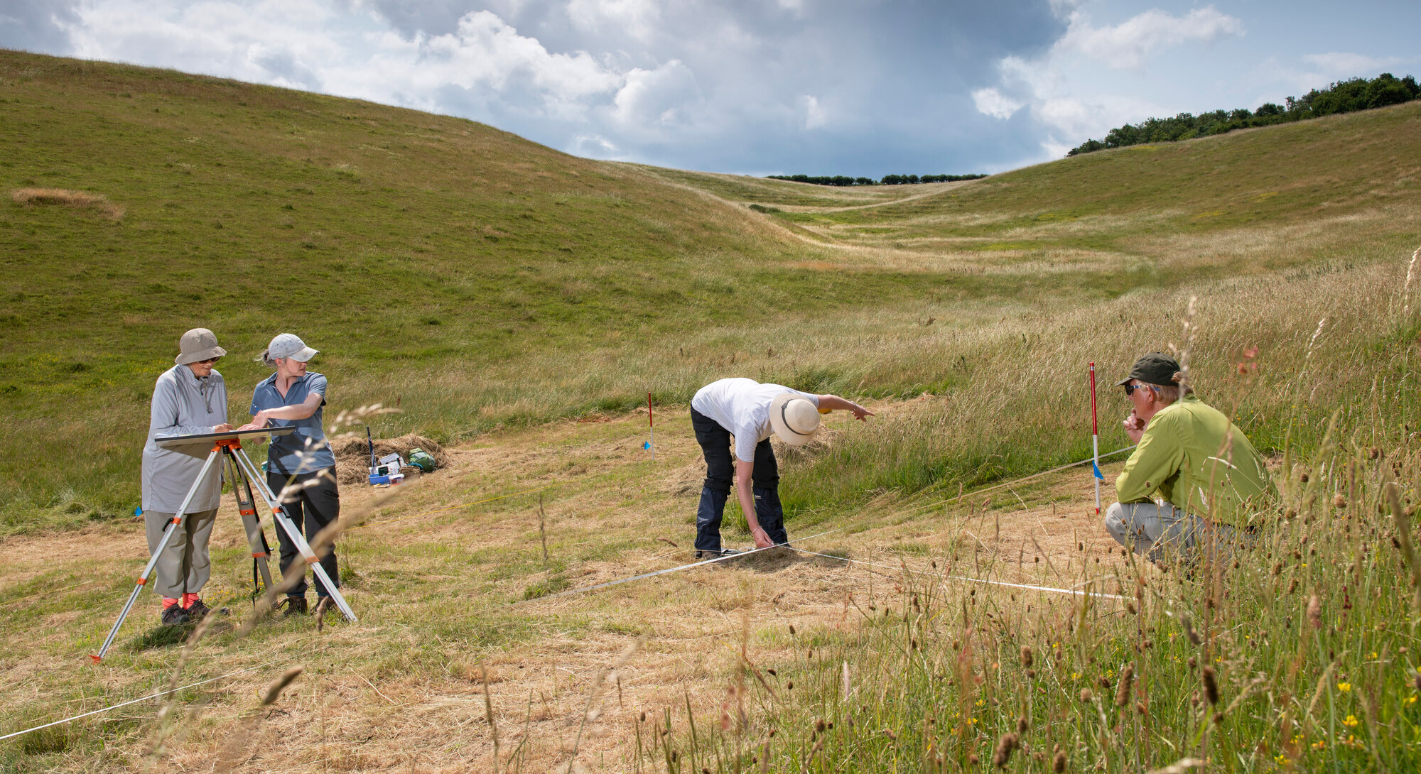A group of four people conducts fieldwork in a grassy landscape, using surveying equipment under a partly cloudy sky, surrounded by rolling hills and natural scenery.