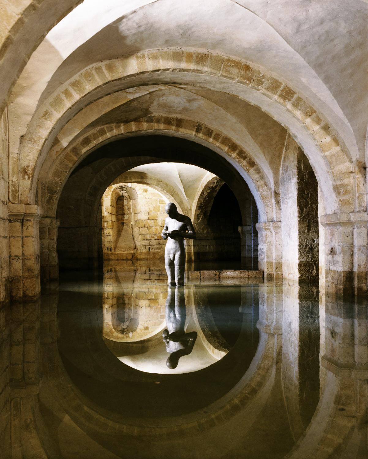 A statue of a human figure stands in flood water up to the knees in a room with vaulted ceilings.