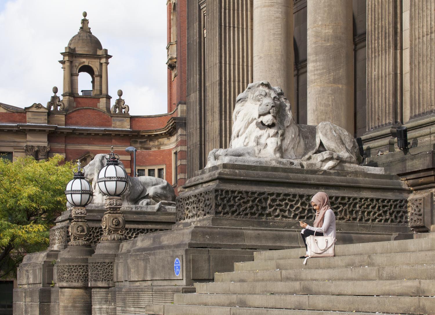 Woman looking at phone on steps to Leeds Town Hall with lion sculpture and columns beyond, view from south east