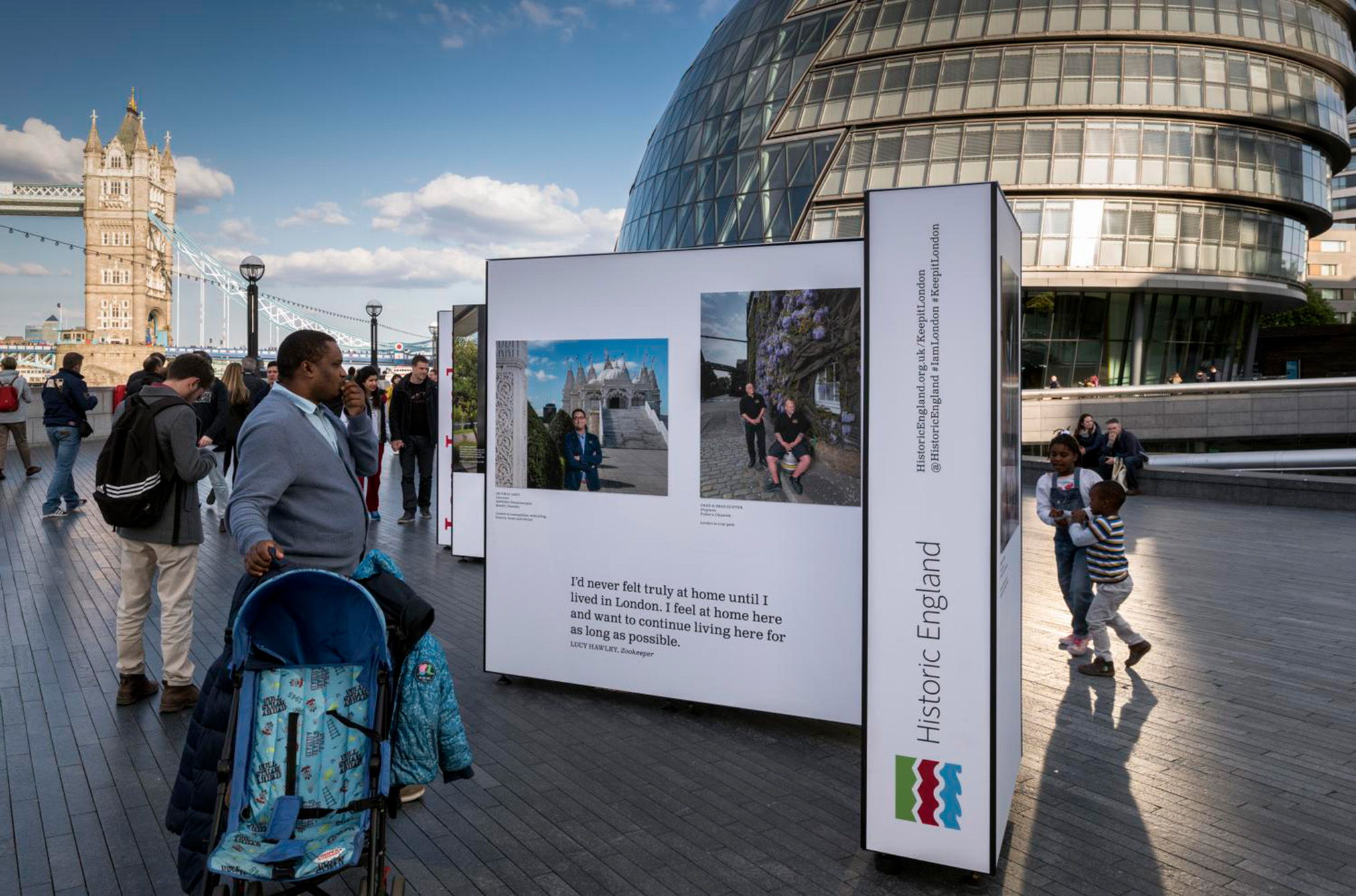 Outdoor exhibition near tower bridge, displaying panels with photos and texts, with people passing by and city hall in the background.