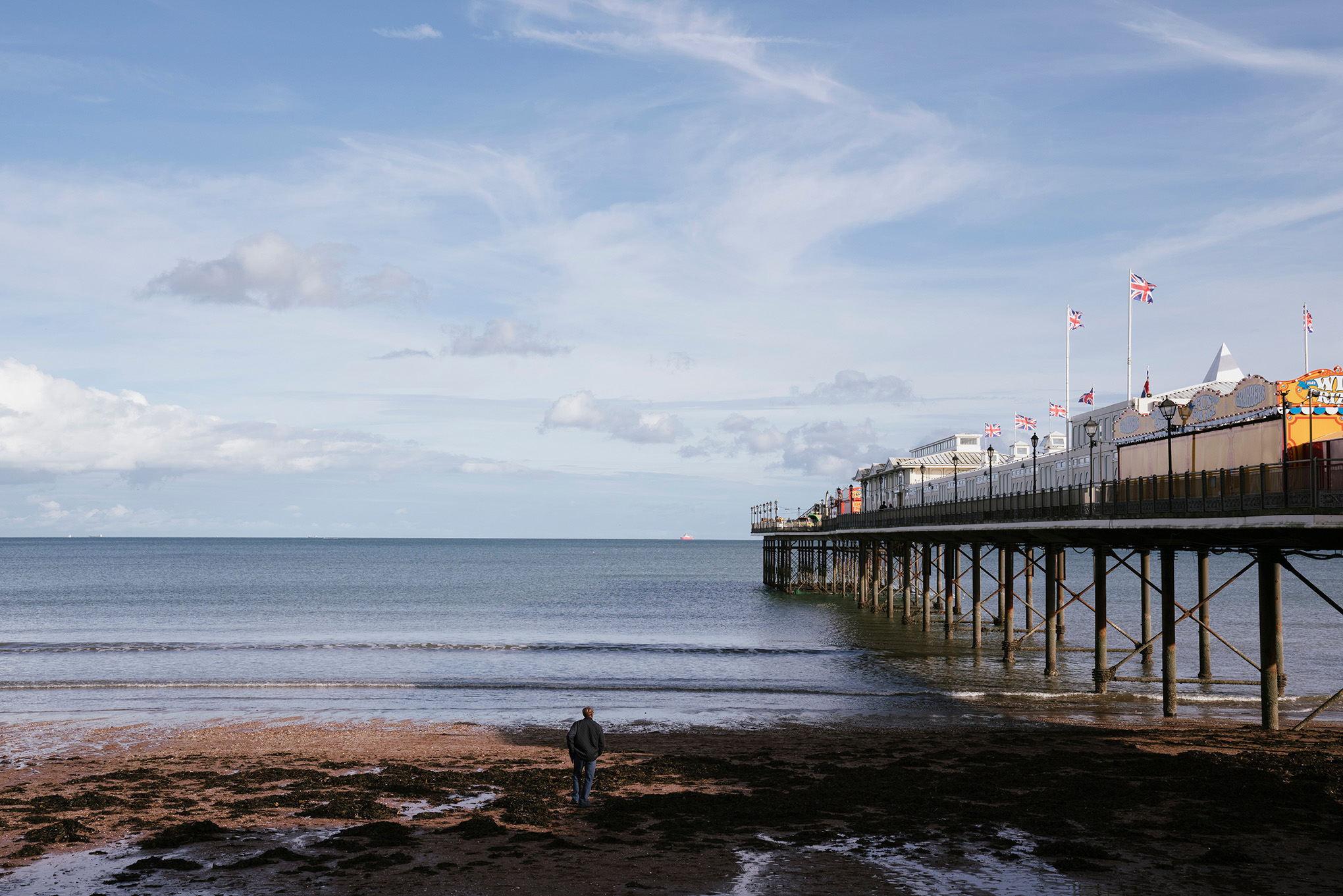 Image of the beach with a pier on right hand side of image and man looking out to sea in the centre.