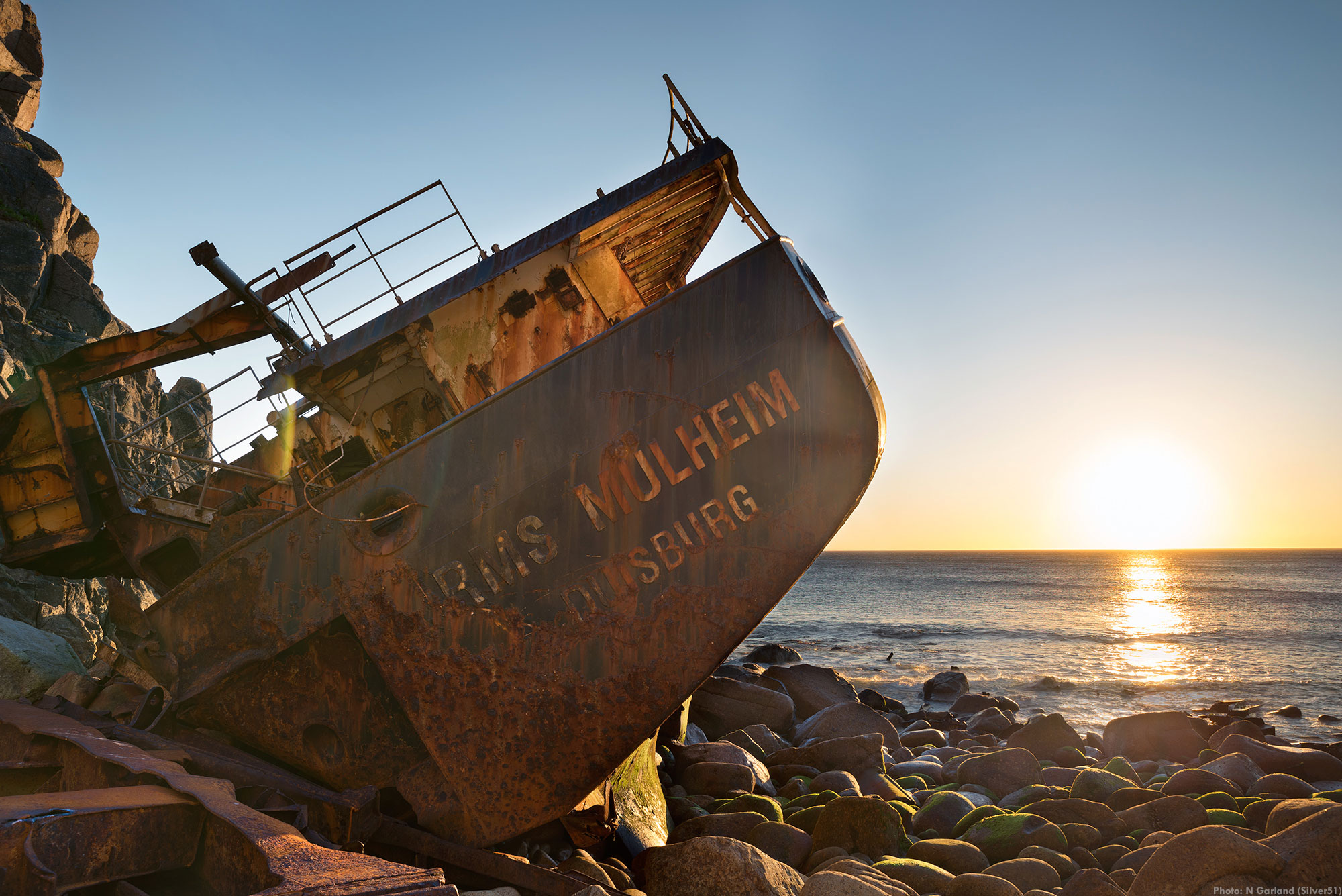 A colour photograph of a wrecked ship on a rocky shore, with the sun setting on the horizon.