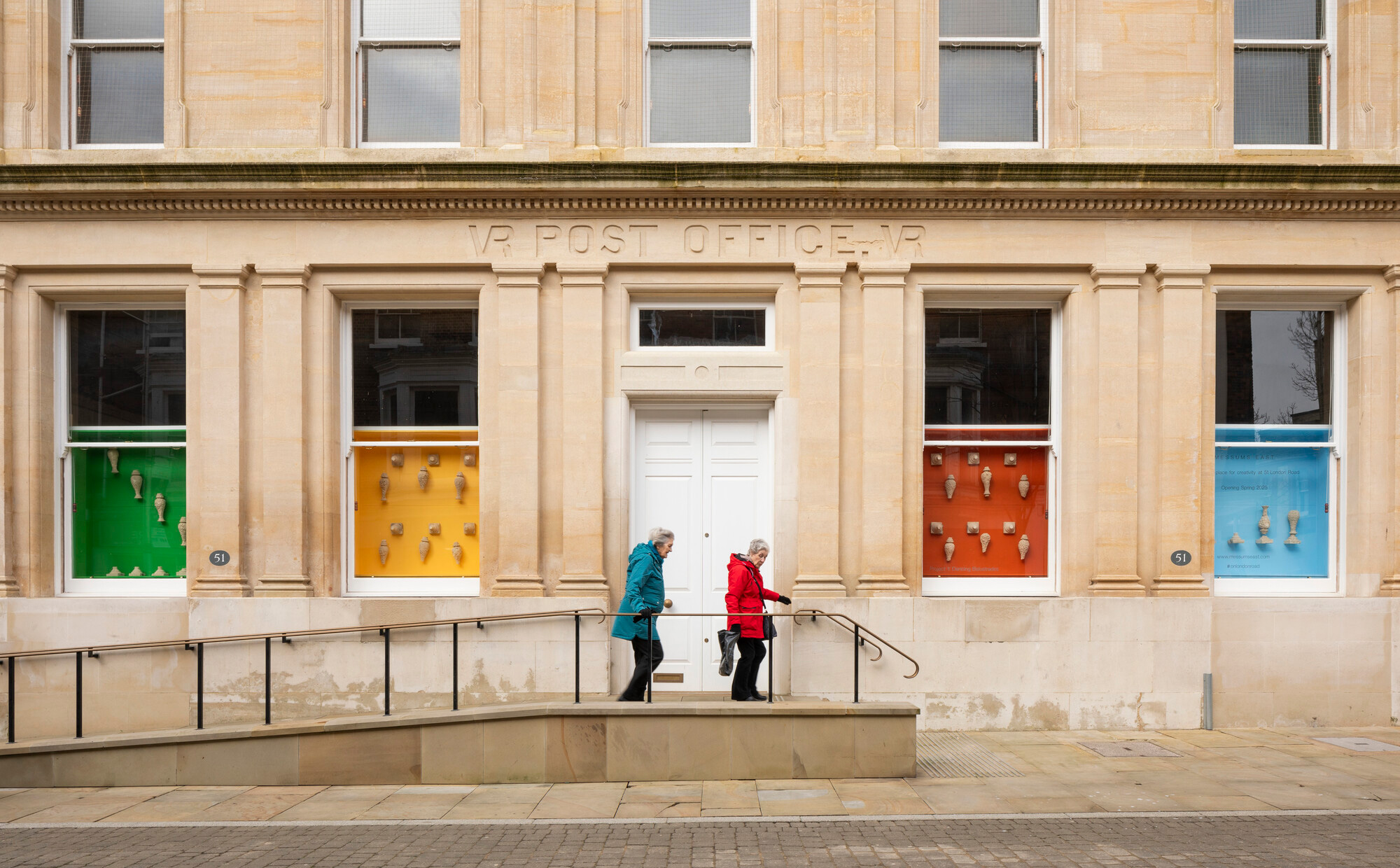 Two people walk on a ramp along the facade of a classic building. Colourful window displays in green, orange, red and blue add a modern touch to the historic post office.