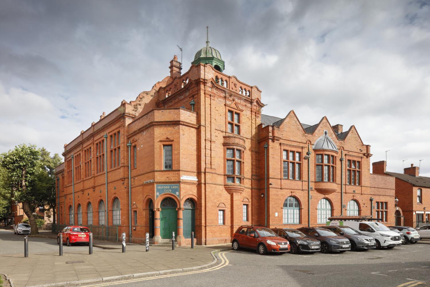 Photograph from road of a red brick building with a green metal tower and large glass windows on the two sides shown. Cars are parked in the carpark in from of the building and a sign over the green front door says Salford Lads Club.