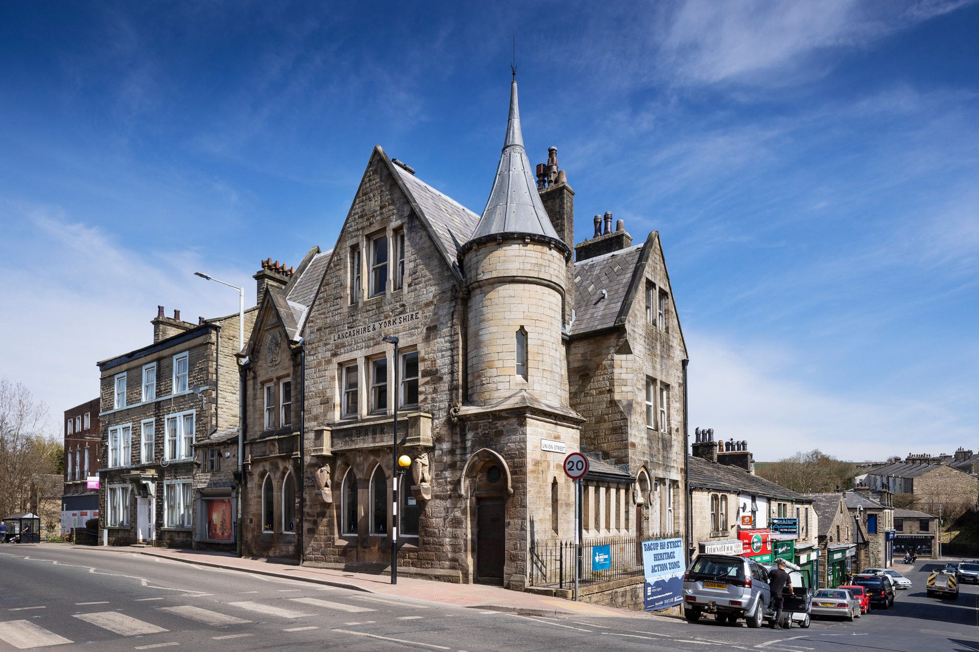 A 3-storey stone building in Scottish baronial style sited on a corner where 2 roads meet. Other buildings on the 2 roads are visible as well as parked cars to the right of the photo.