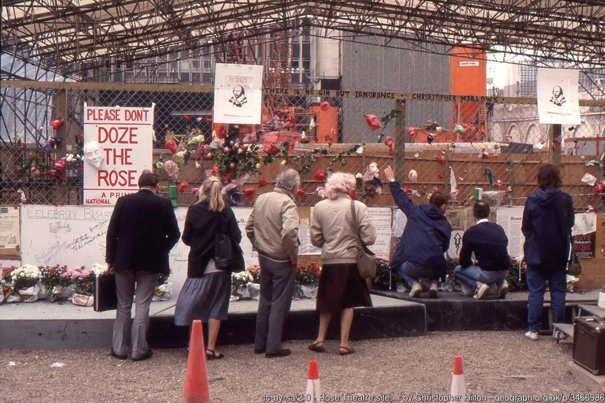 A photograph of seven members of the public looking at the excavation site of the Rose Theatre in London, behind a chain link fence.