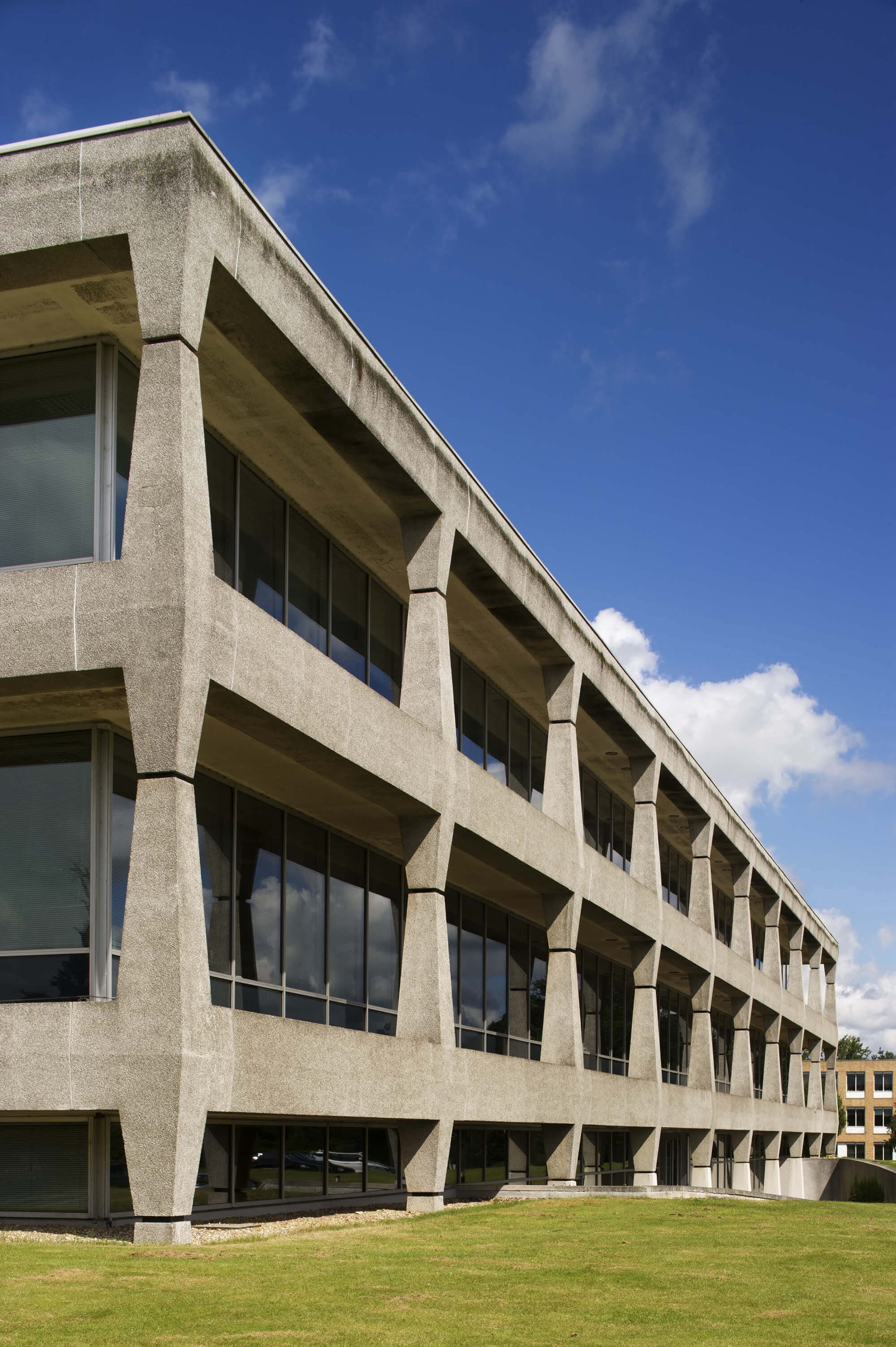 Three storey grey concrete modern building with grass lawn in front and blue skies.