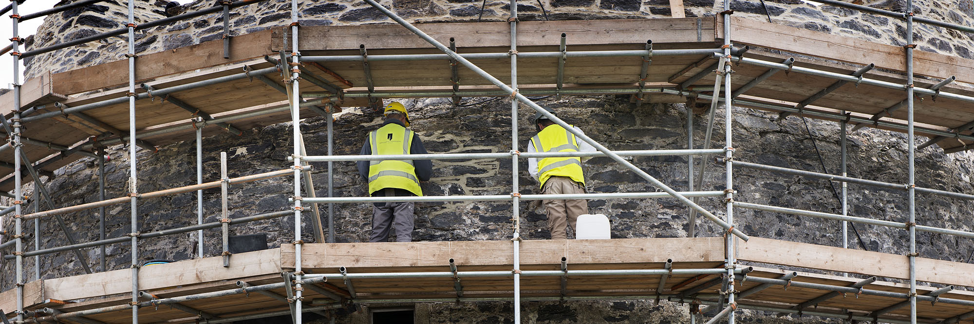 A photograph from behind of two individuals within scaffolding working on a stone wall.