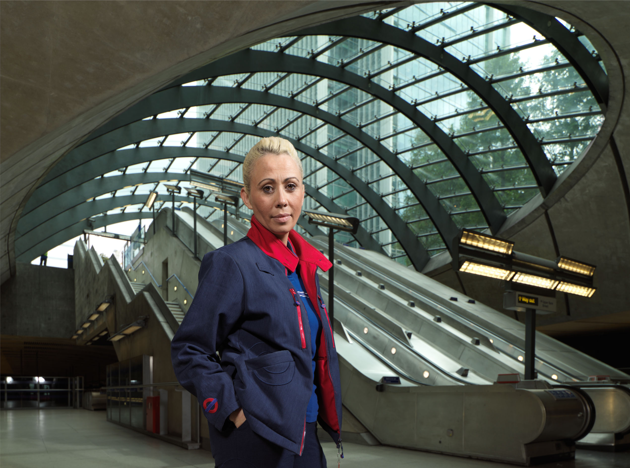 Female London Underground employee standing at the bottom of escalators at Canary Wharf station.