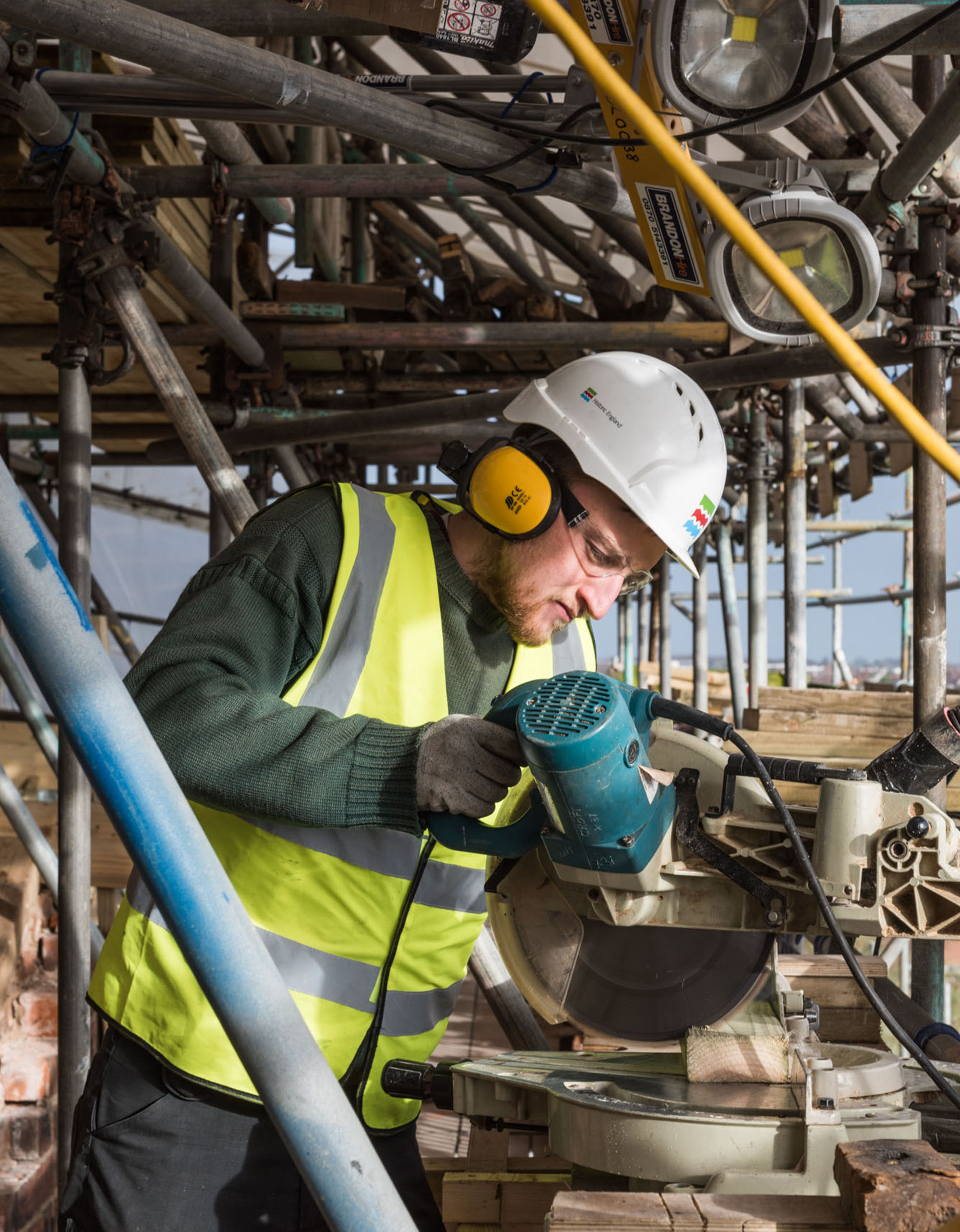 Prince’s Foundation trainee wearing hard hat, safety glasses and ear defenders cutting a piece of wood