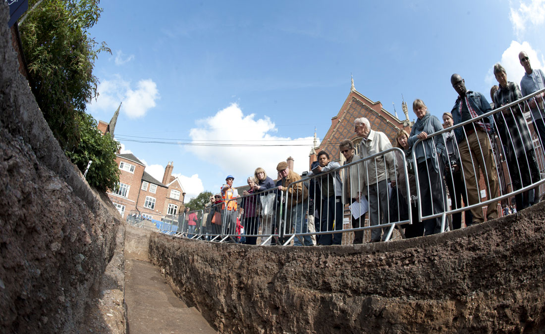 Members of the public peering over fencing into an archaeological trench, photographed from the bottom of the trench looking up.