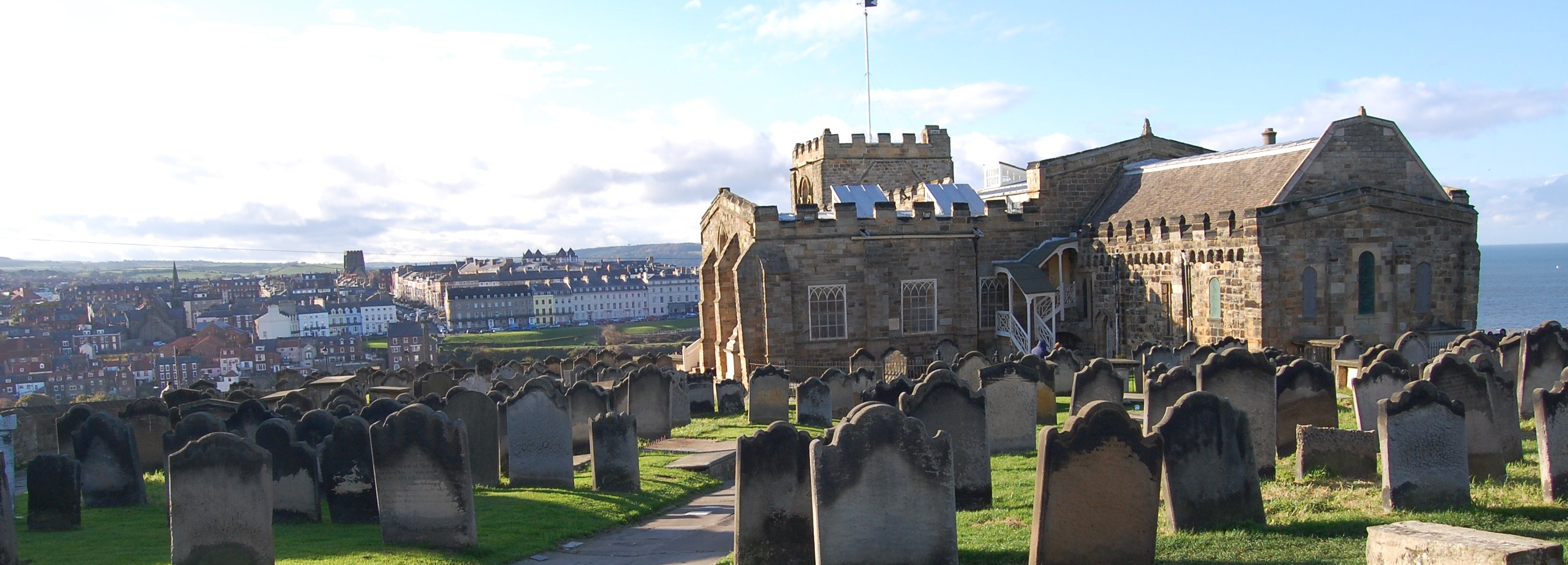 A photographic of a historic church underneath a blue cloudy sky, with gravestones in the foreground and the seaside town of Whitby in the background.