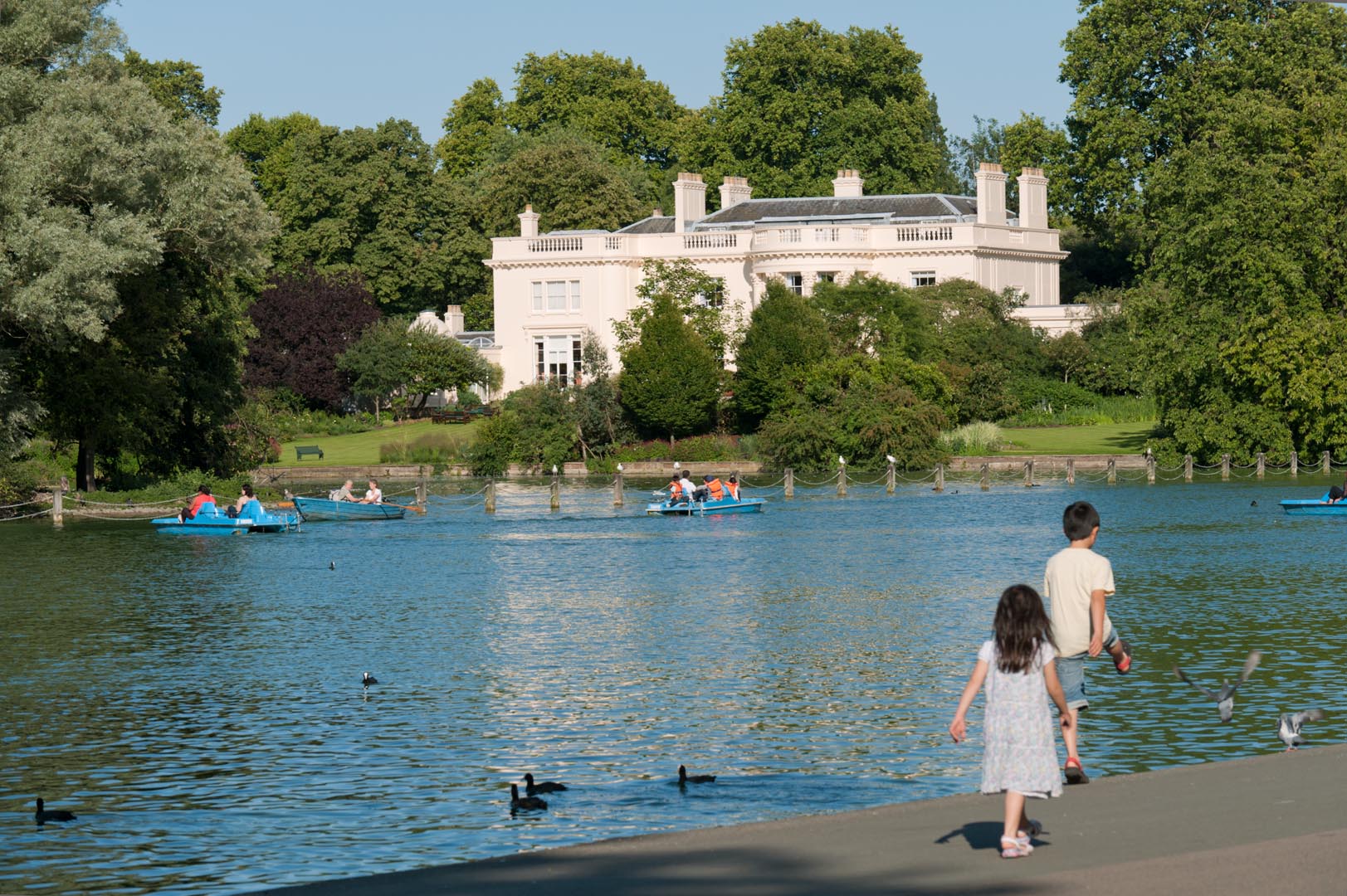 The Holme, children walking along waterfront with multiple boats in it. The house and parks in the background