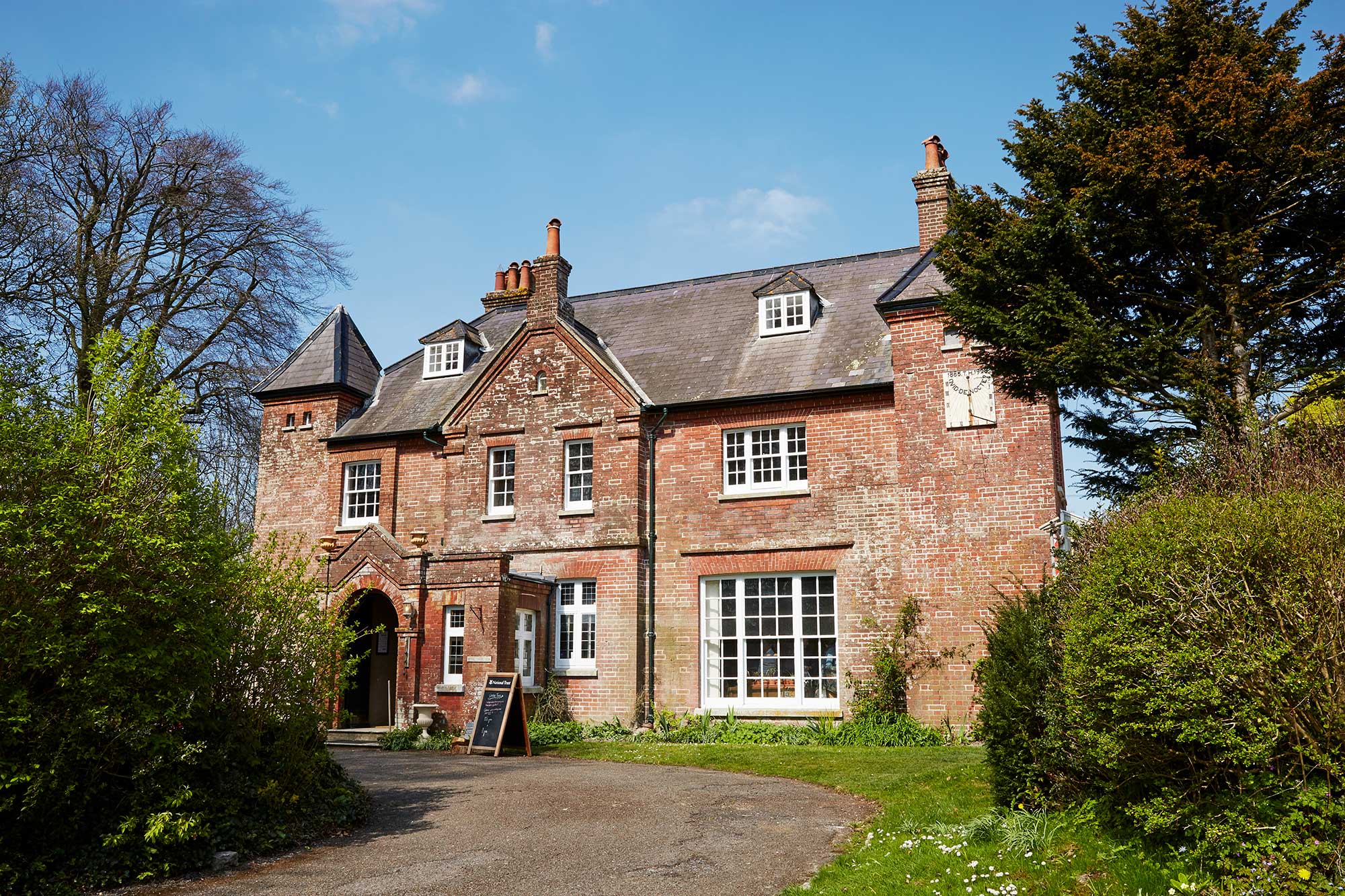 Red brick house with sash windows surrounded by trees and bushes.