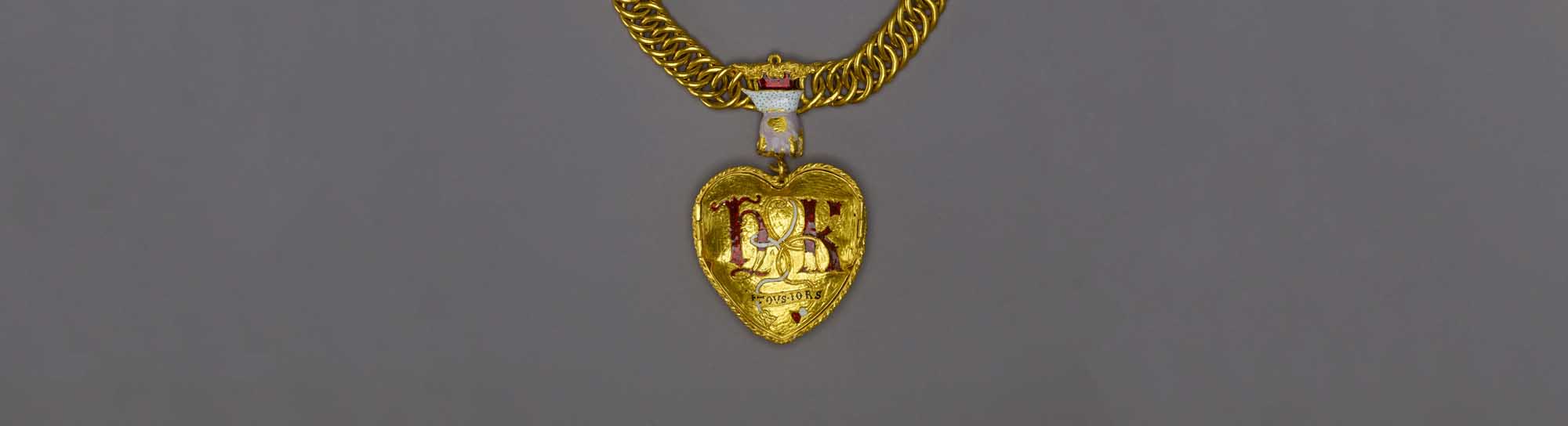 A large, heart-shaped gold pendant on a thick gold chain, suspended from a hand-shaped ornamentation, and detailed with a rose and pomegranate. 