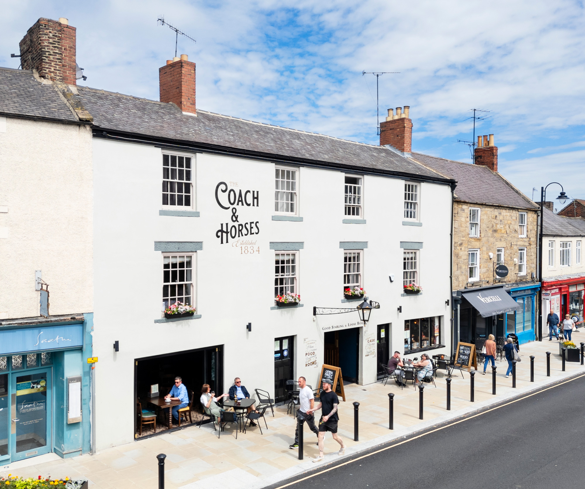 Pub with some customers settled at outdoor seeting on pavement outside, and pedestrians passing by.