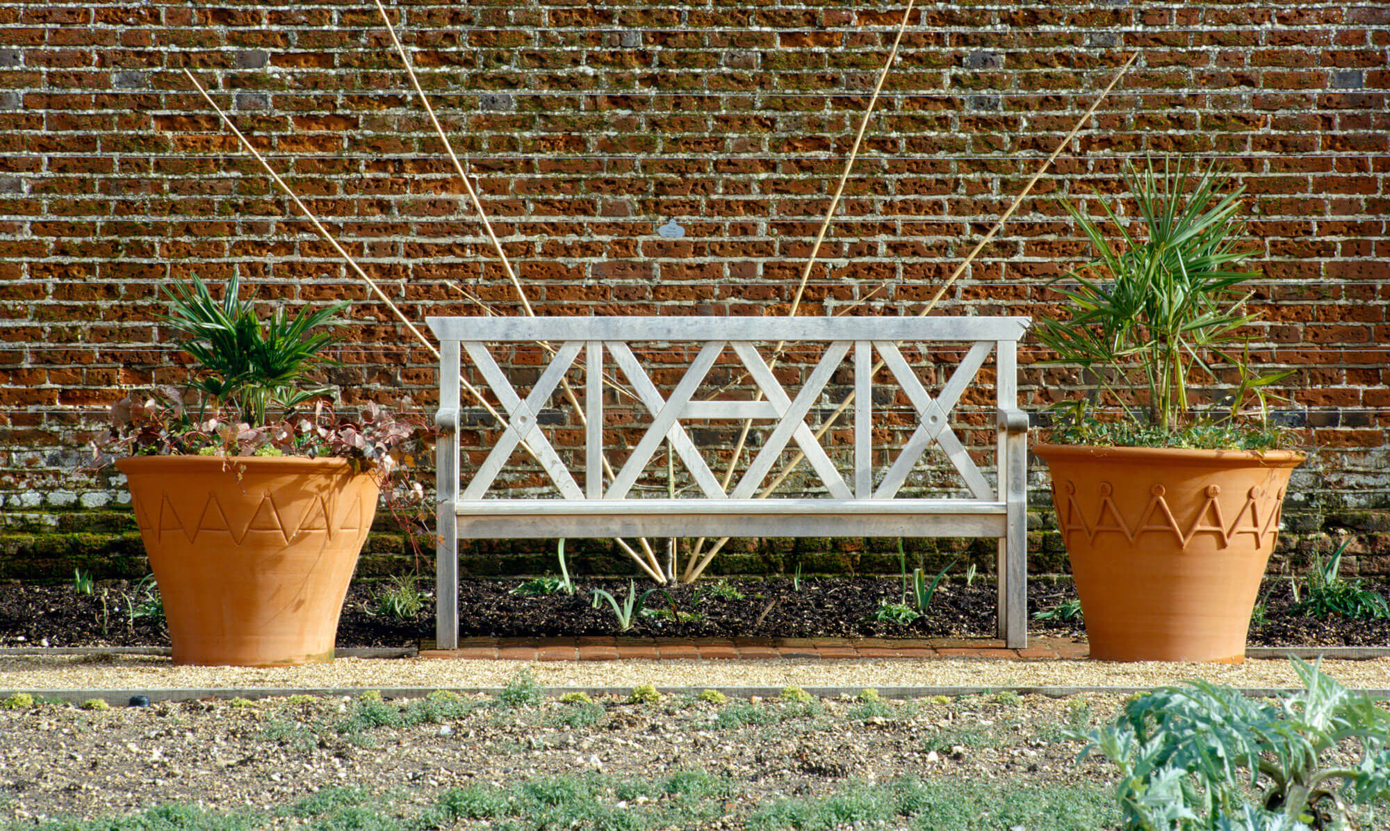 A wooden bench with a geometric design sits between two large terracotta pots, adorned with plants, against a textured brick wall.